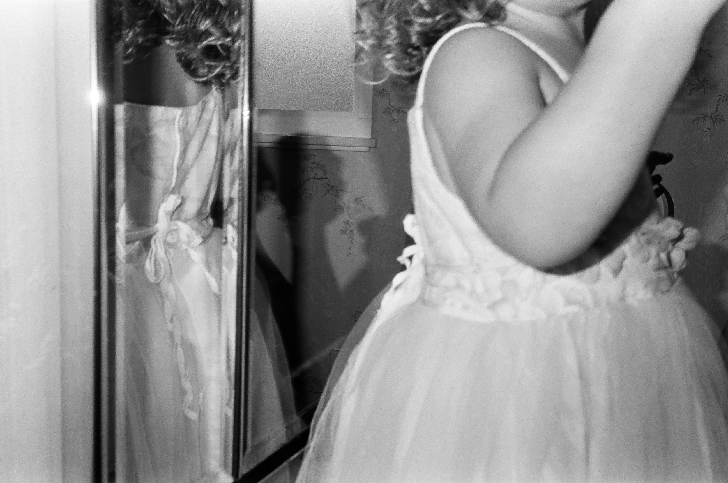 A girl wearing a light-colored dress with ruffles, standing in front of a mirror, taking a selfie in a room with patterned wallpaper. Oakland family photos in film. 