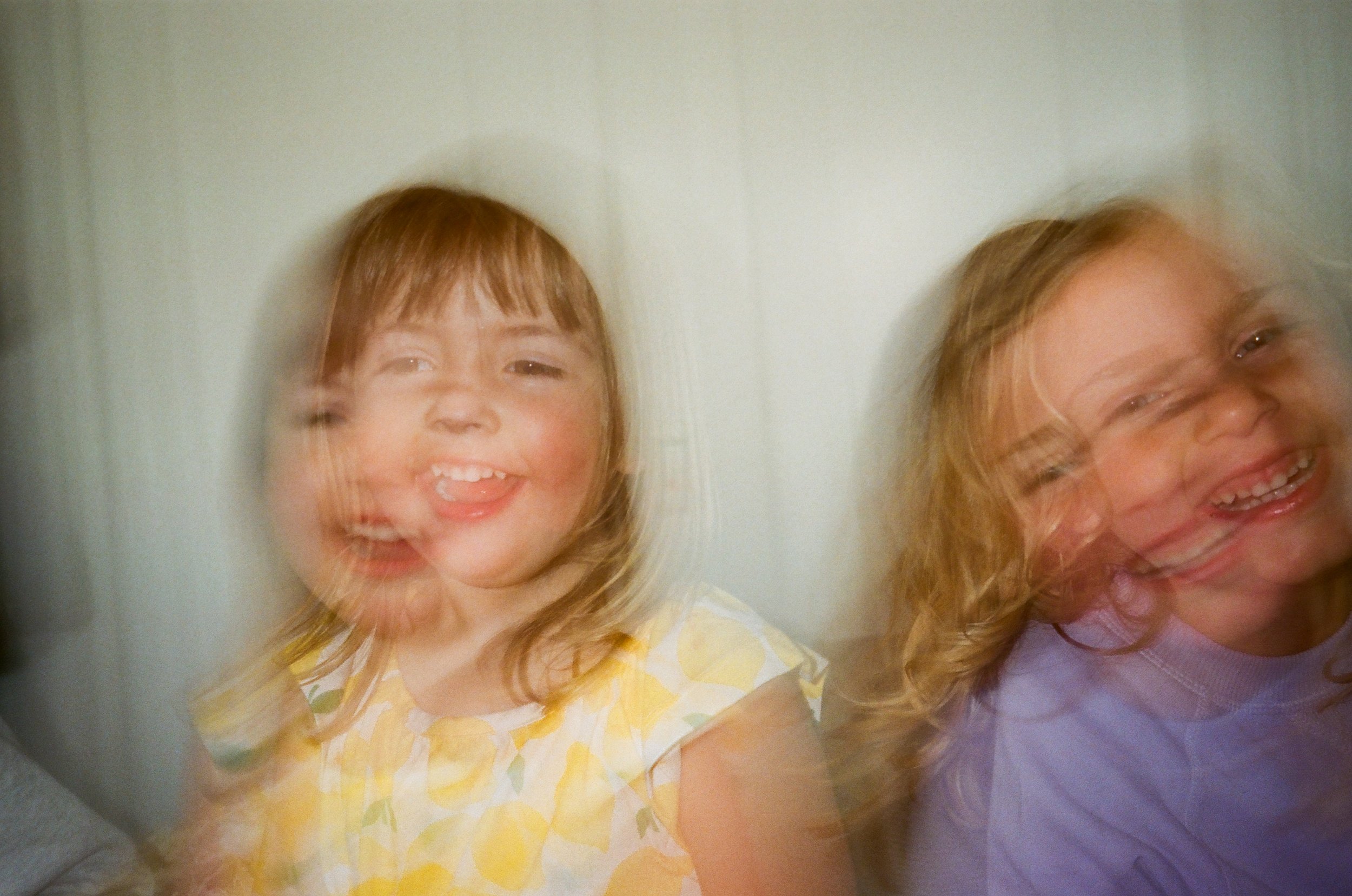 Two young girls with blonde hair smiling and appearing to move or blur, sitting indoors against a plain light-colored wall. Slow shutter in film, San Francisco moving photos. 