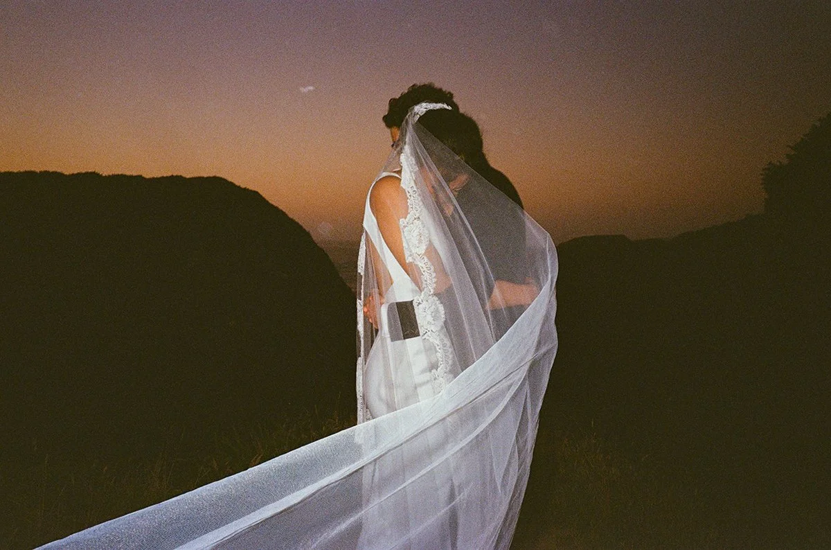 A couple dressed in wedding attire, standing close together in an outdoor setting during dusk, with silhouetted hills in the background and serving a romantic atmosphere.