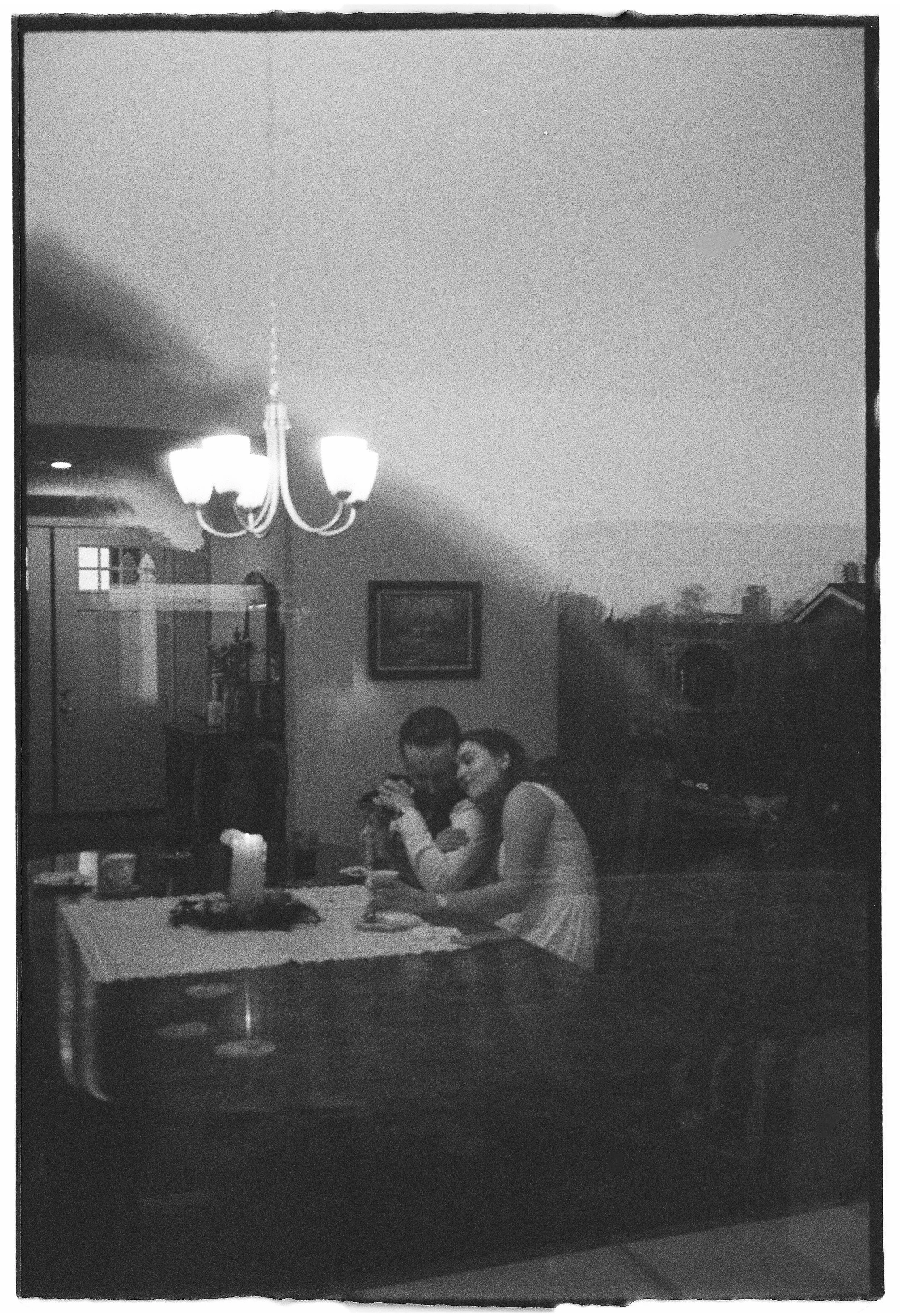 A black and white photo of a couple sitting at a dining table inside, seen through a window. The reflection of a chandelier and a candle on the table are visible, with a painting on the wall outside and the silhouette of houses in the background.