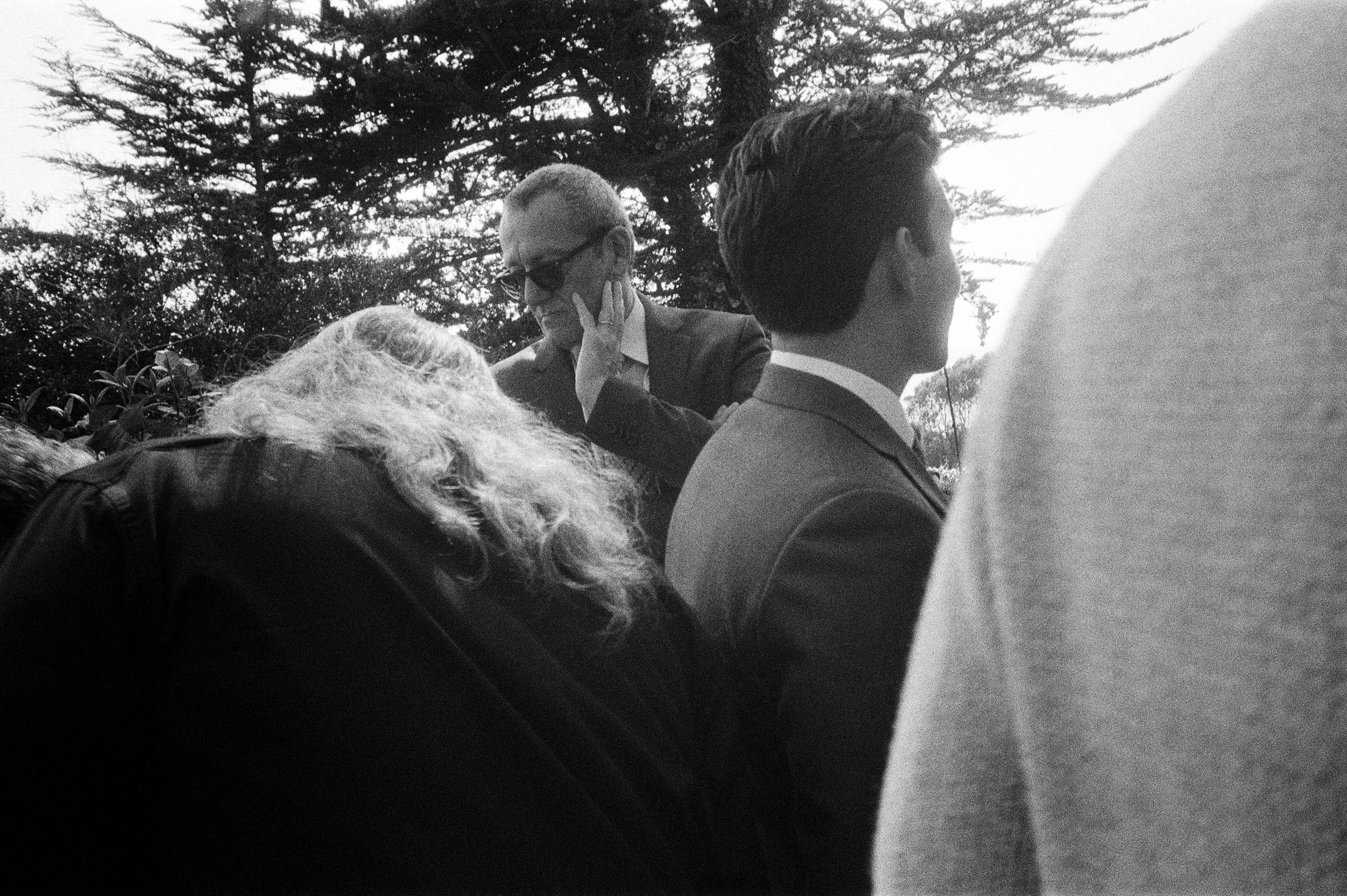 A group of people outdoors, some talking and one person wearing sunglasses and a suit, with trees in the background. Switzer Farm Westport, California 