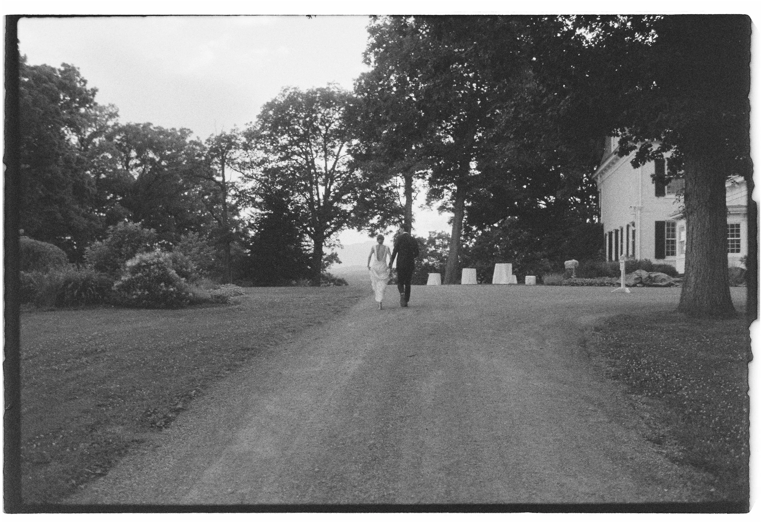 A black-and-white photo of a couple walking hand in hand on a dirt path in front of a large house, surrounded by tall trees and lush bushes. Hudson Valley wedding in film. 
