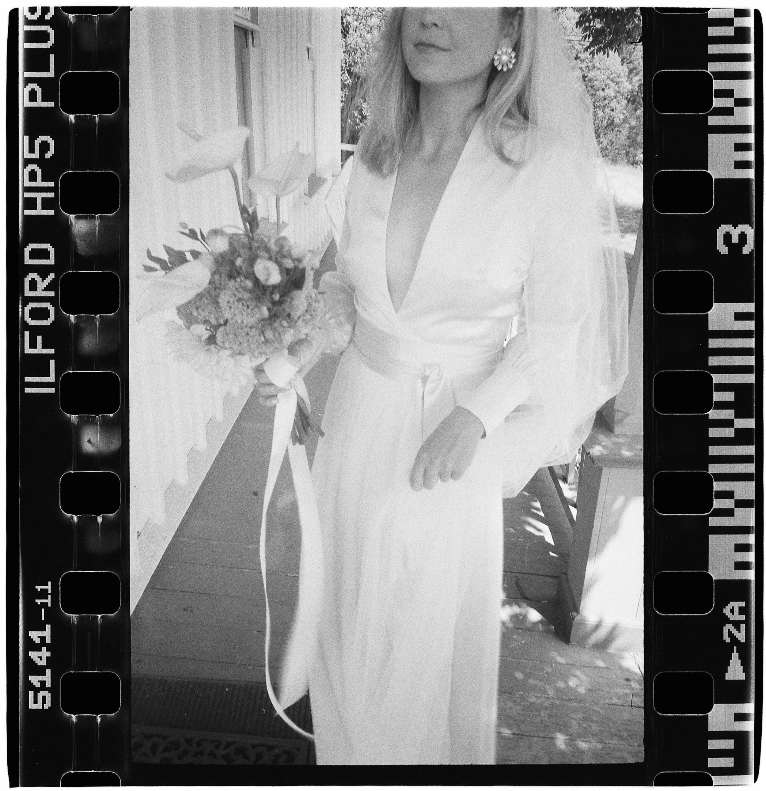 A woman in a satin wedding dress holding a bouquet of flowers, standing outdoors on a wooden porch at Spring Ranch in Mendocino California. Film. 
