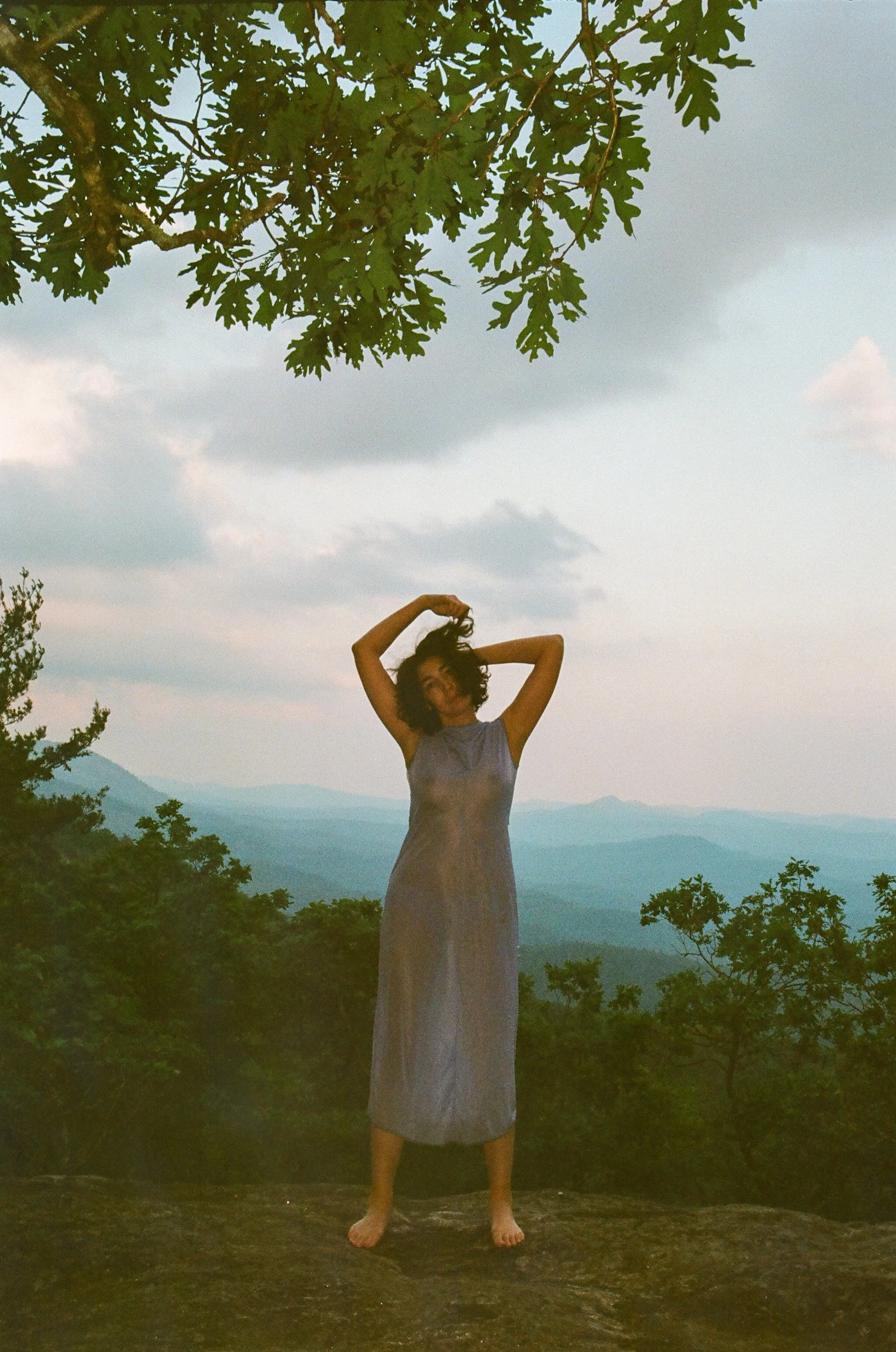 A woman standing barefoot on a rock at sunset, with mountains and a cloudy sky in the background. She is wearing a sleeveless, long dress and has curly hair, with her arms raised above her head. Blueridge mountains. 