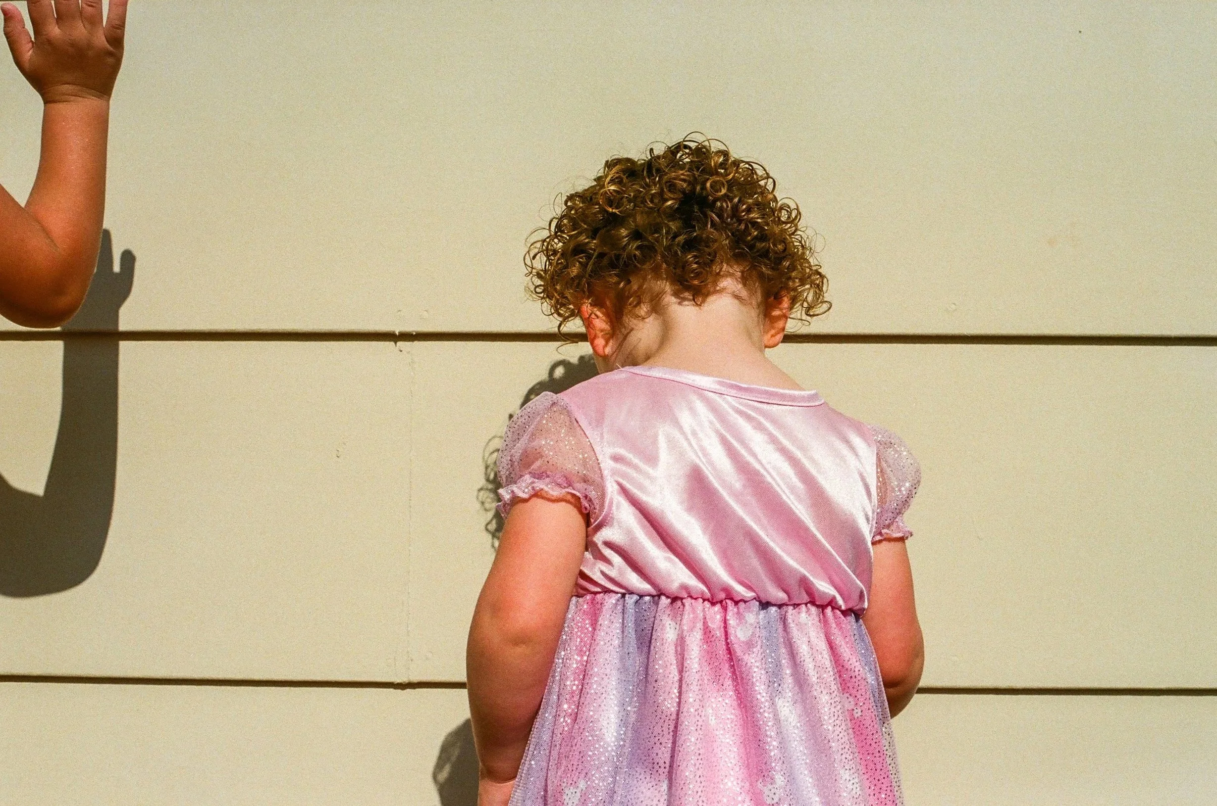 A young girl with curly hair wearing a pink dress stands with her back to the camera, facing a beige wall. Part of another child's arm is visible on the left side of the image. Portland film family photos. 