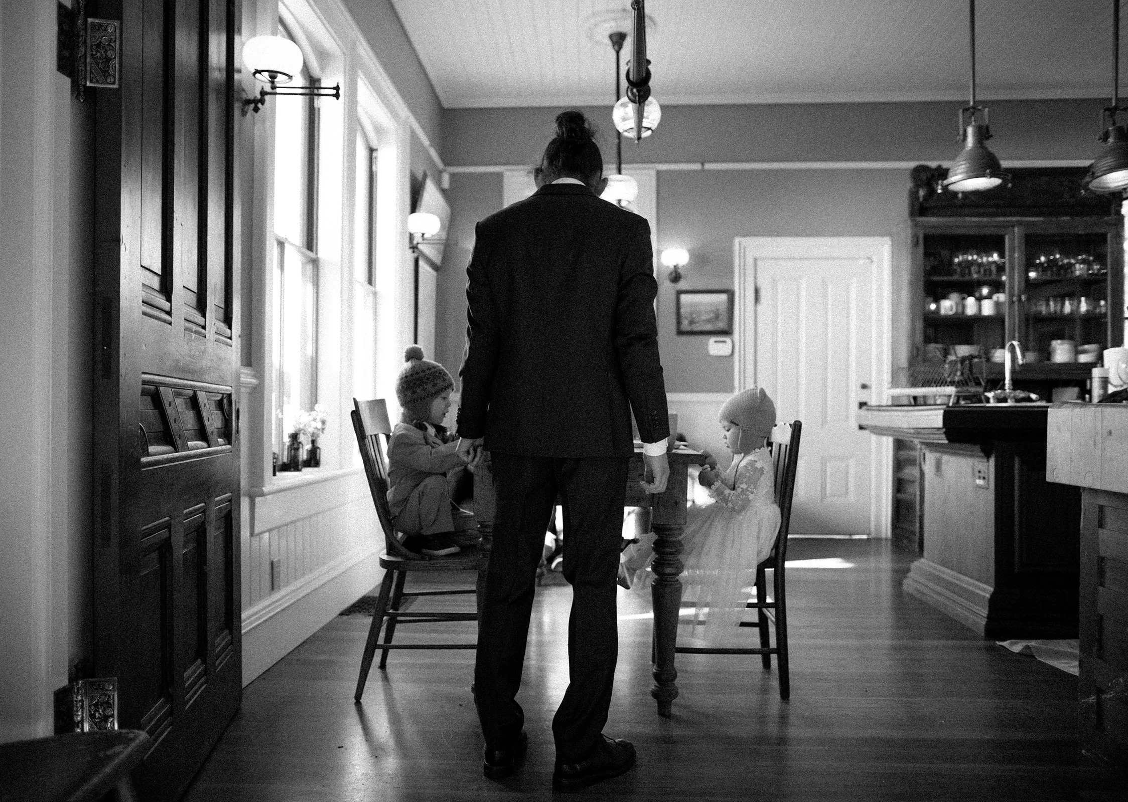A person in a suit standing in a dining room with two children, one dressed casually and the other in a dress and hat, seated at a table, with a kitchen in the background.