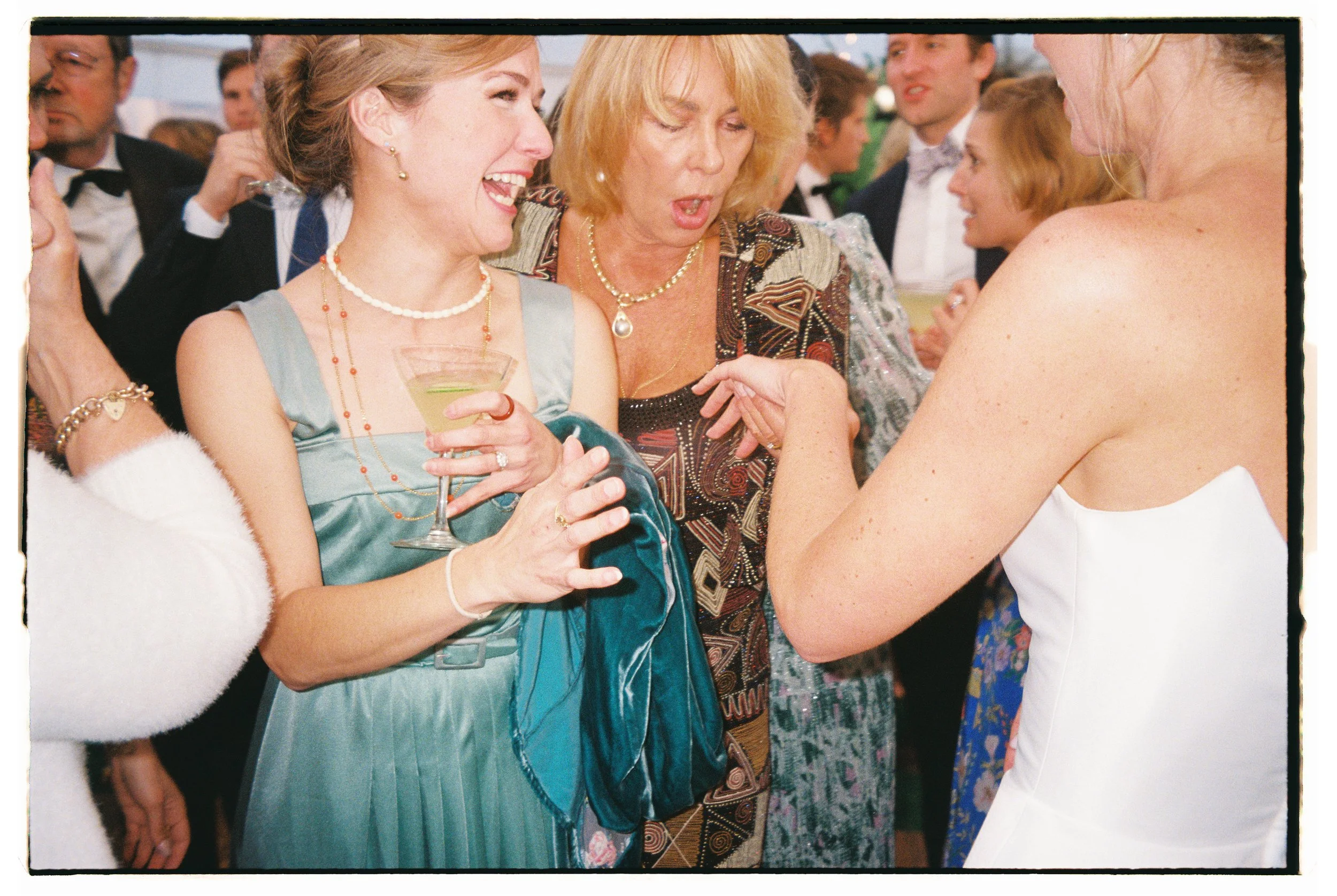 People at a lively party, socializing, with women in elegant dresses and men in tuxedos, holding drinks and engaging in conversation.