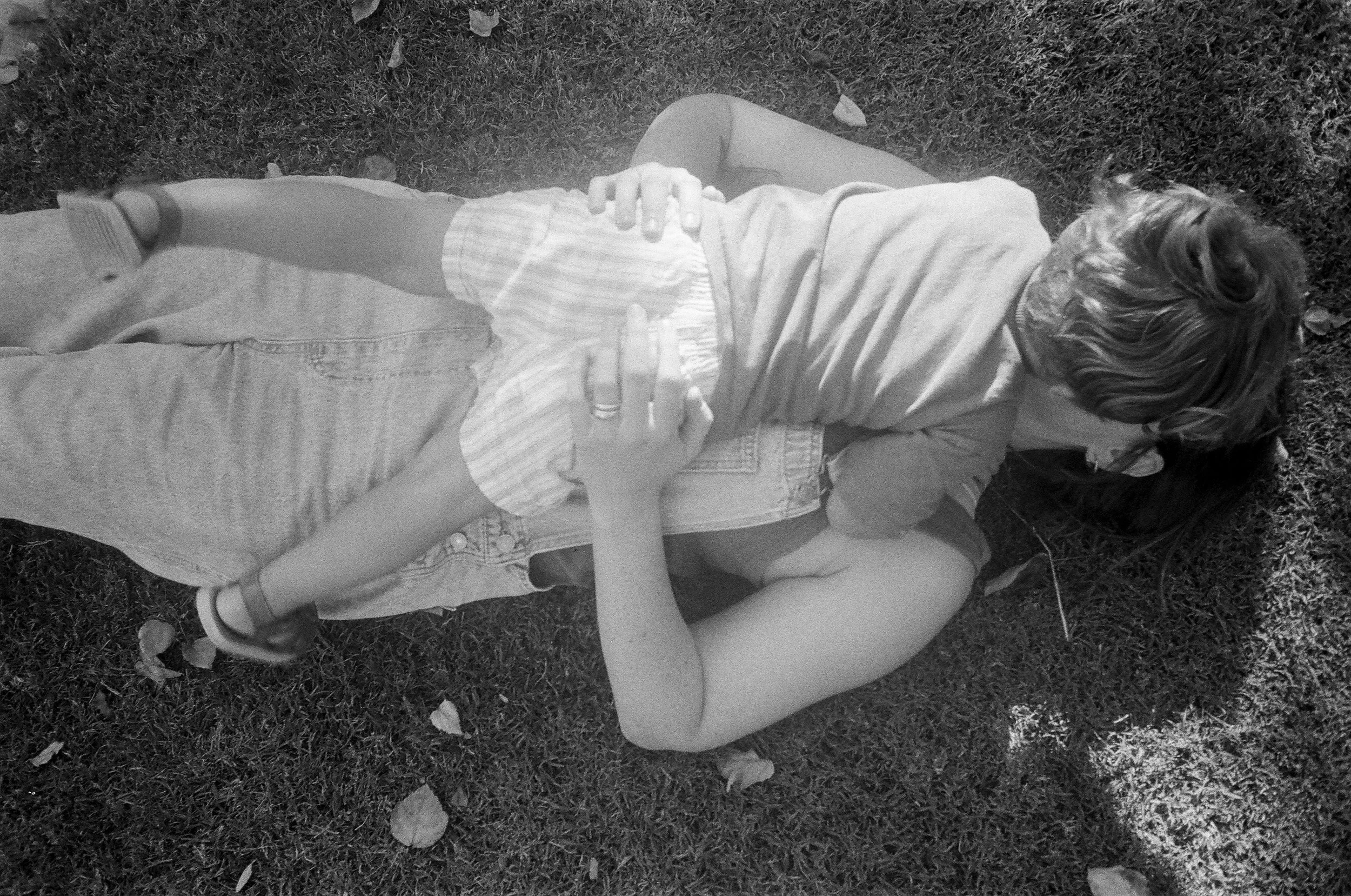 A woman and a young girl lying on grass, embracing each other, with the woman's hand on the girl's back. The scene is outdoor, with some fallen leaves around. Film Palo Alto, California multi generational family photos. 