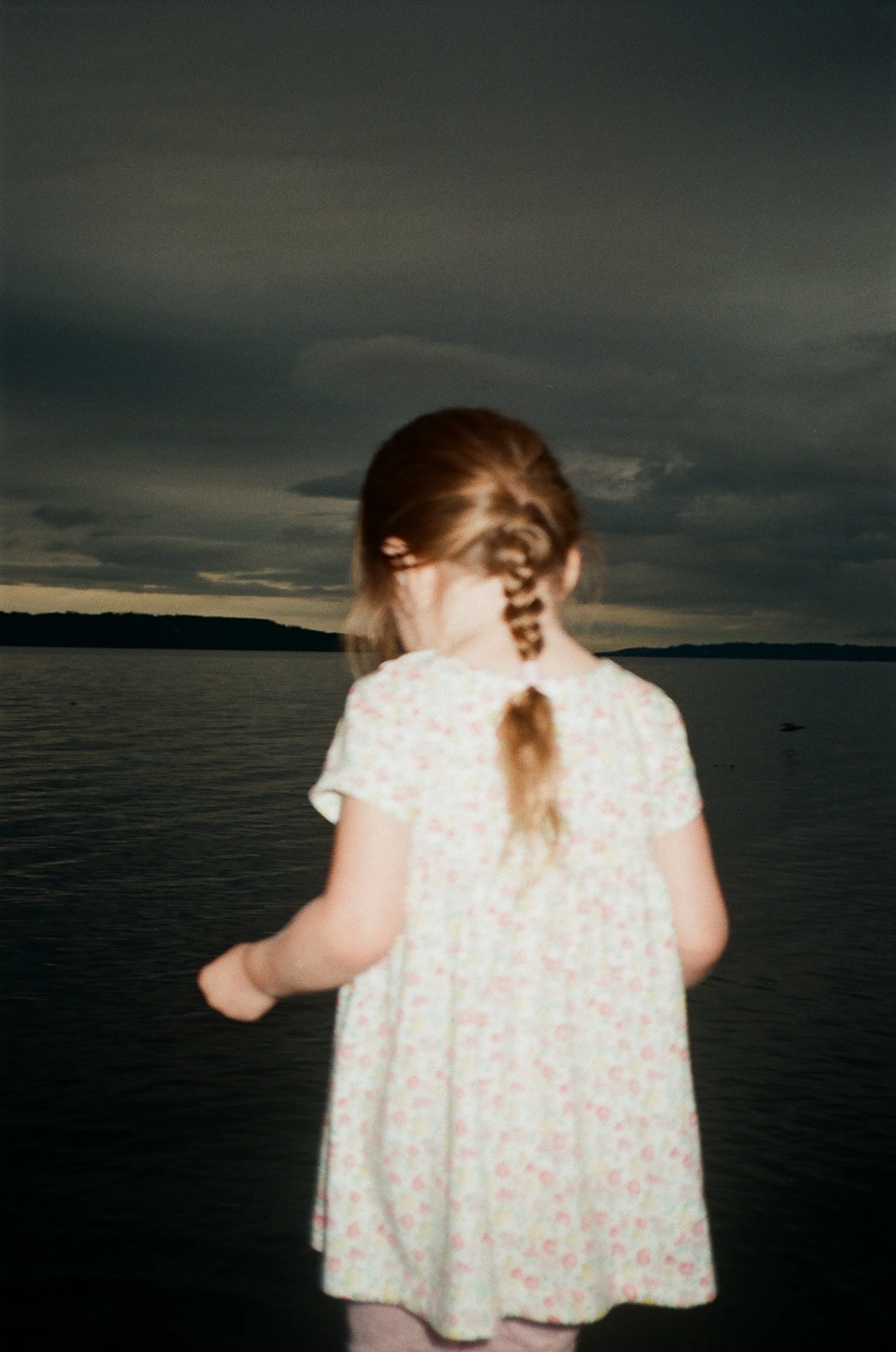 A young girl with braided hair stands by a body of water, facing away from the camera, under a cloudy, dark sky.