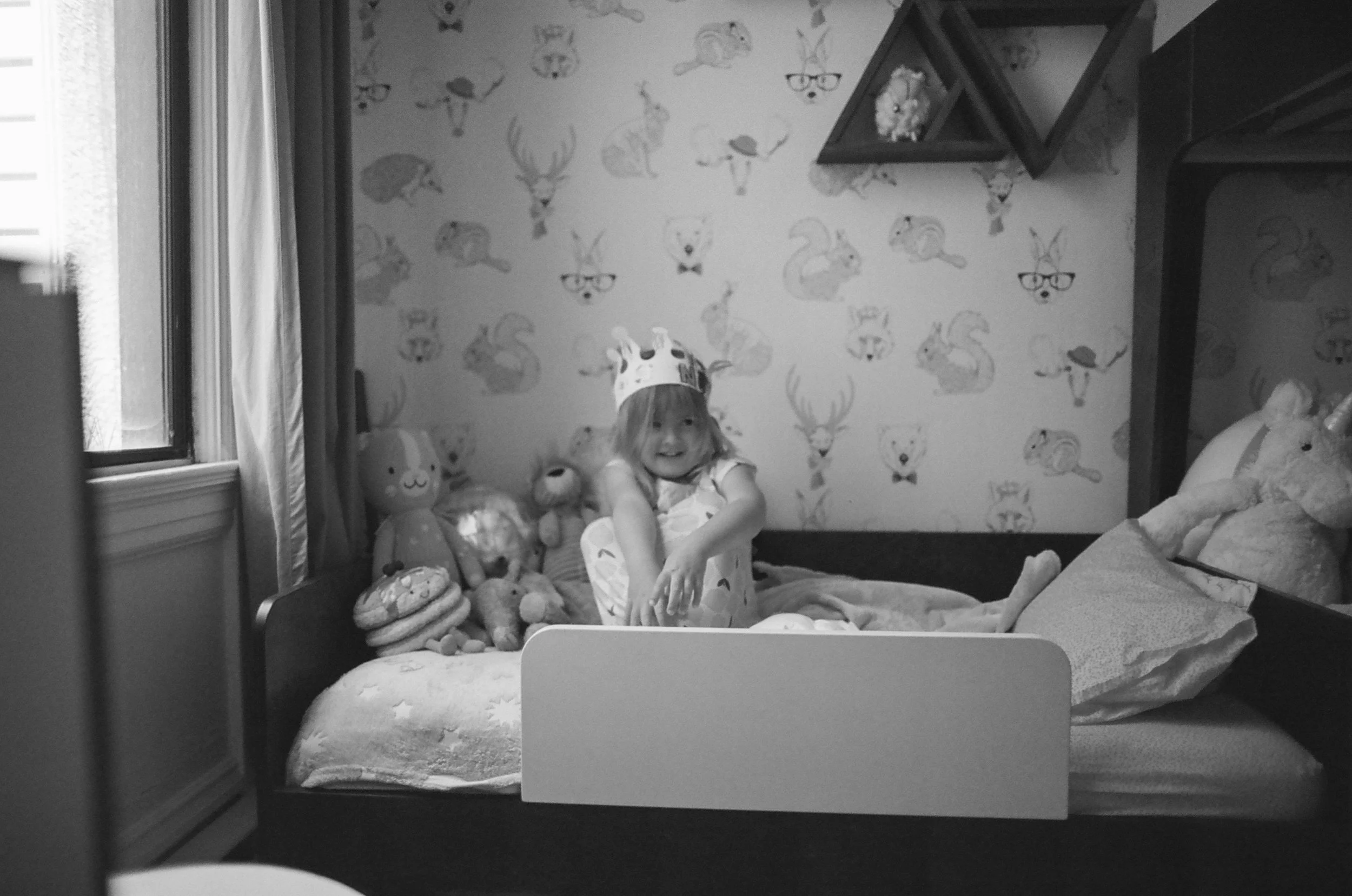 A young girl wearing a paper crown and a dress, sitting on a bed surrounded by stuffed animals, smiling at the camera in a child's bedroom.