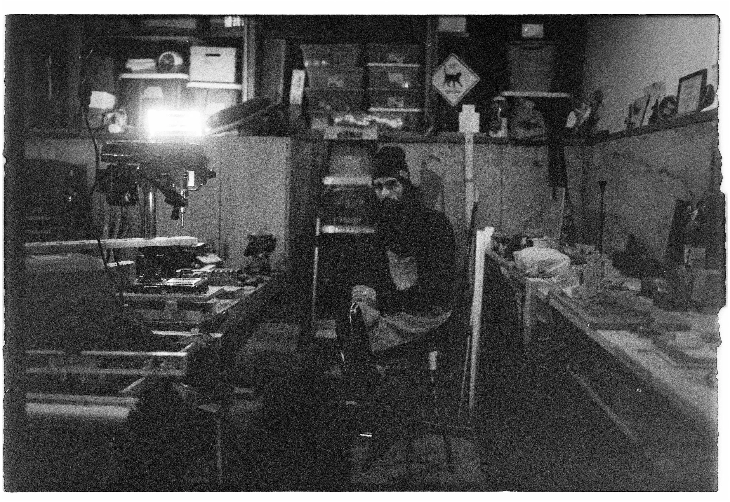 A man sitting in a cluttered workspace, surrounded by tools, boxes, and electronic equipment in black and white. Film Tacoma Washington. 