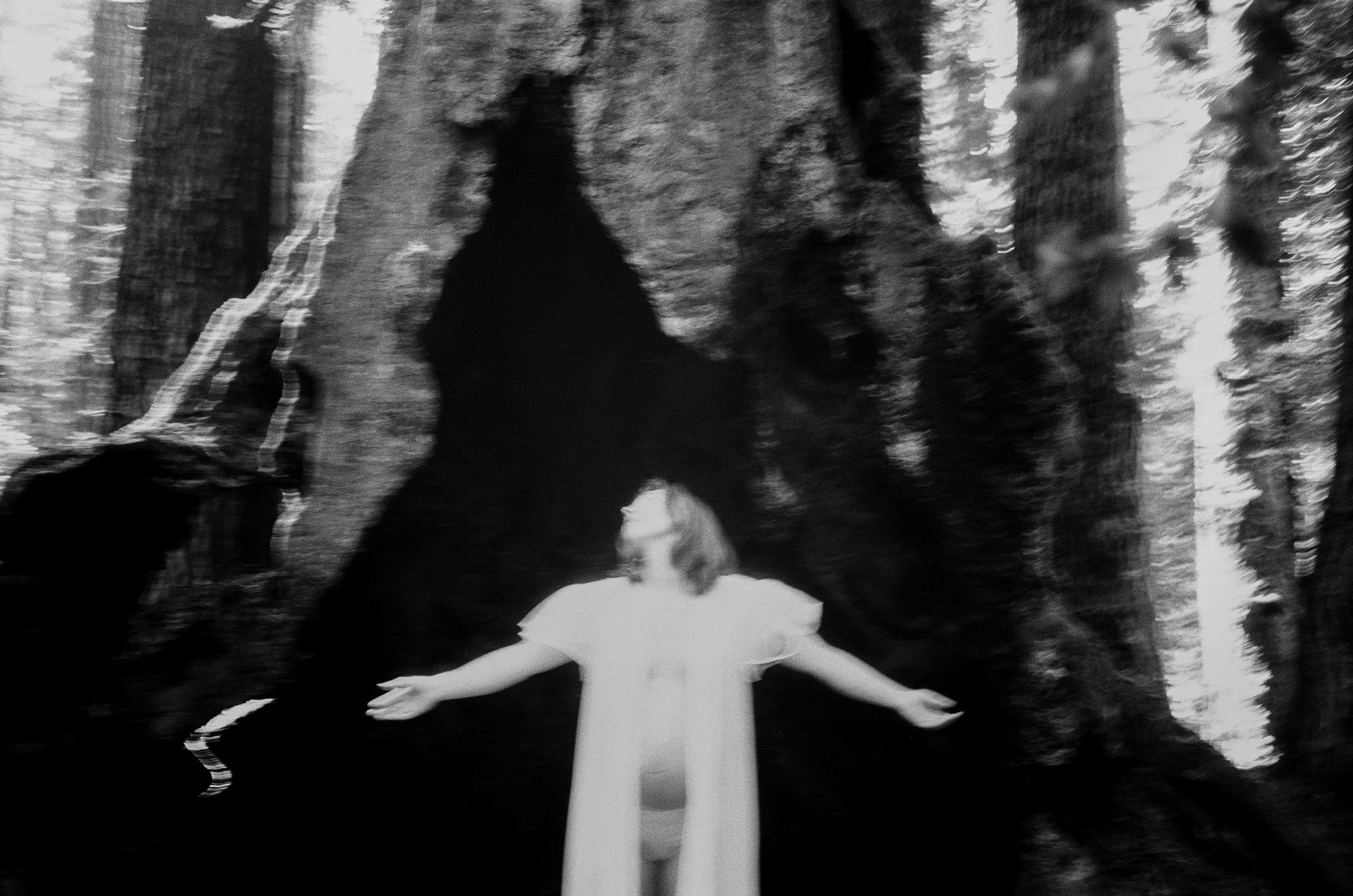 Black and white photo of a woman standing with arms outstretched in front of a large rock formation in a forest. Maternity photos redwoods Mendocino, the farm. 