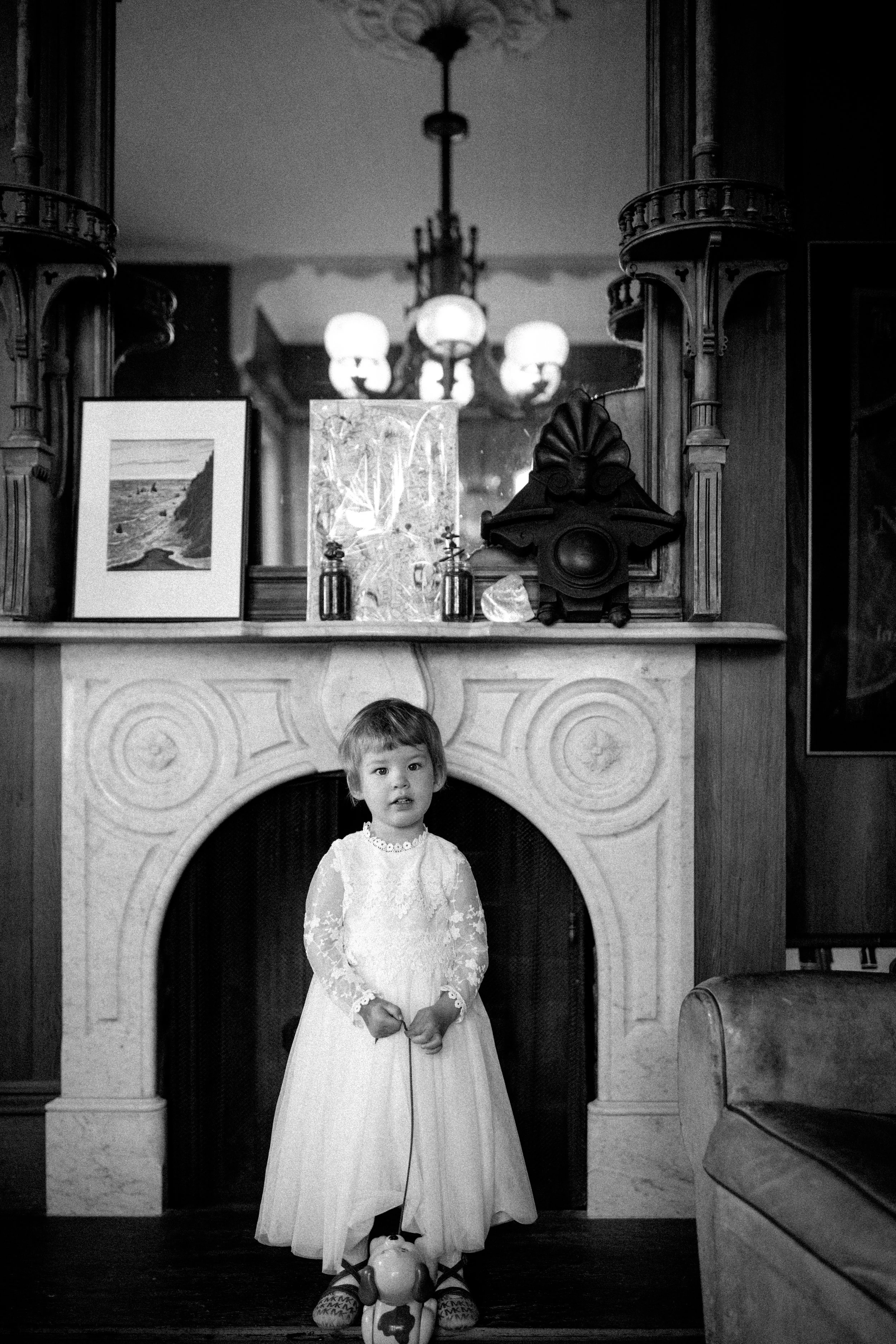 A young girl in a white lace dress standing in front of a fireplace, holding a toy pump. The background features a large mirror, a framed picture, and decorative objects, with a chandelier hanging from the ceiling. Switzer Farm, Mendocino. 
