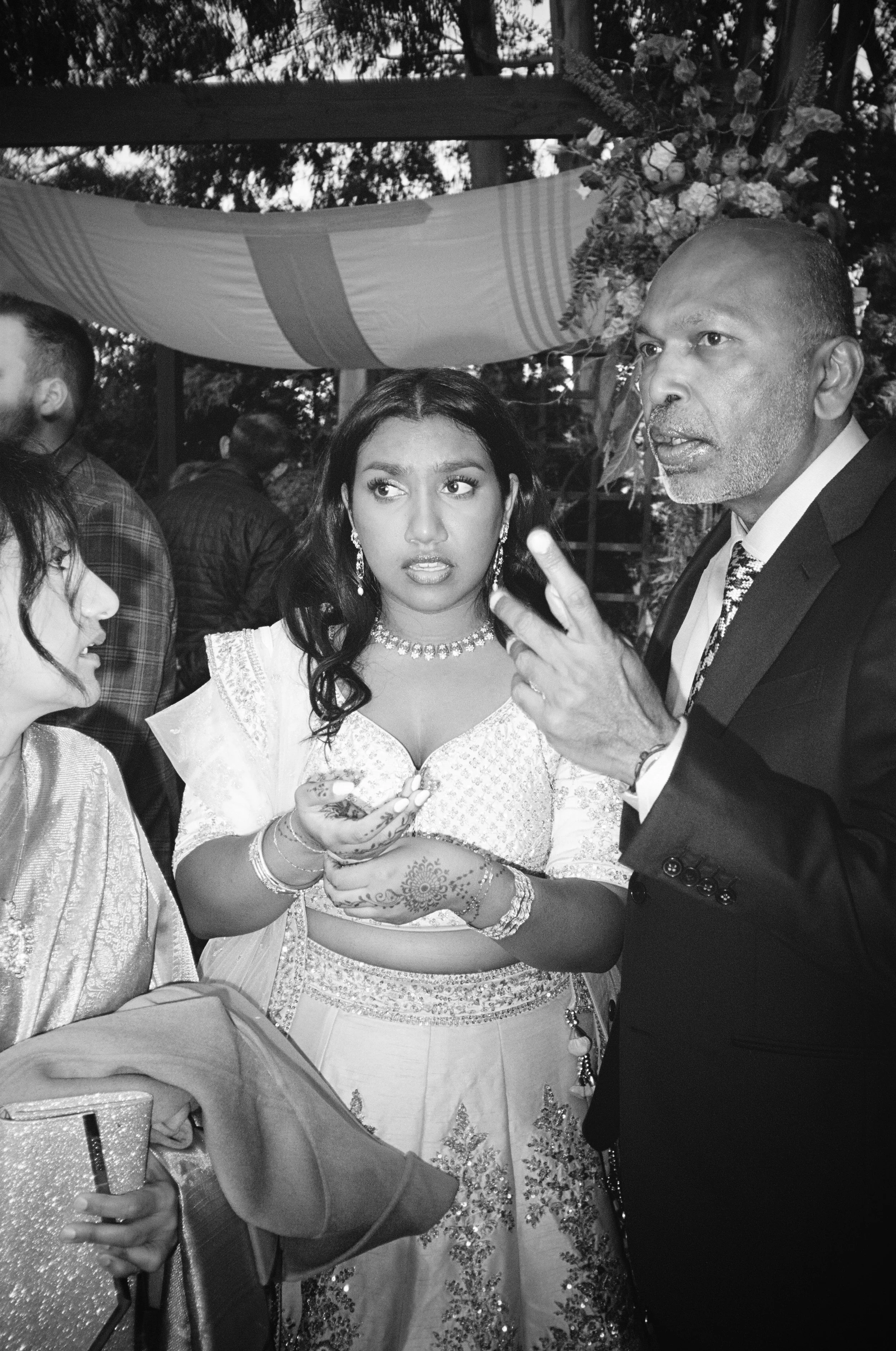 A black-and-white photo of a man in a suit having a conversation with two women dressed in traditional Indian attire, standing outdoors under a decorated canopy with flowers. Indian wedding golden gate club film.  