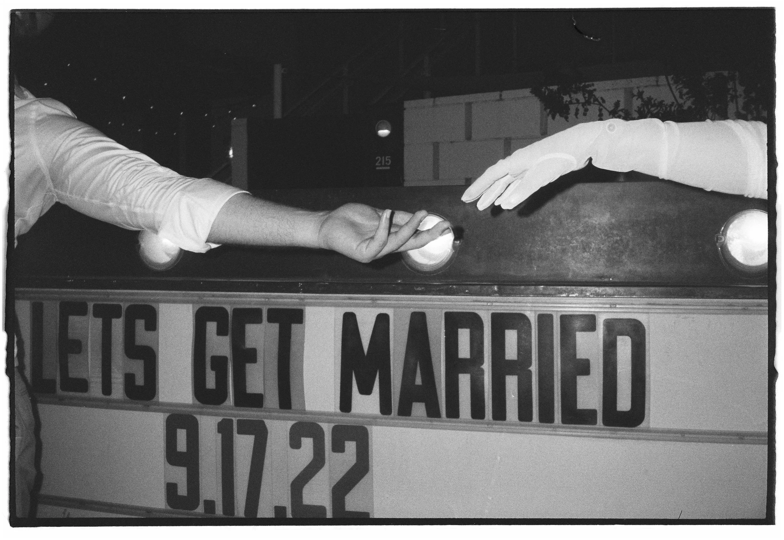 A black and white film photograph of a couple reaching, with the bride's hand wearing long white gloves over a sign that reads "Let's Get Married" and a date, September 22. Ace hotel, Palm Springs. 