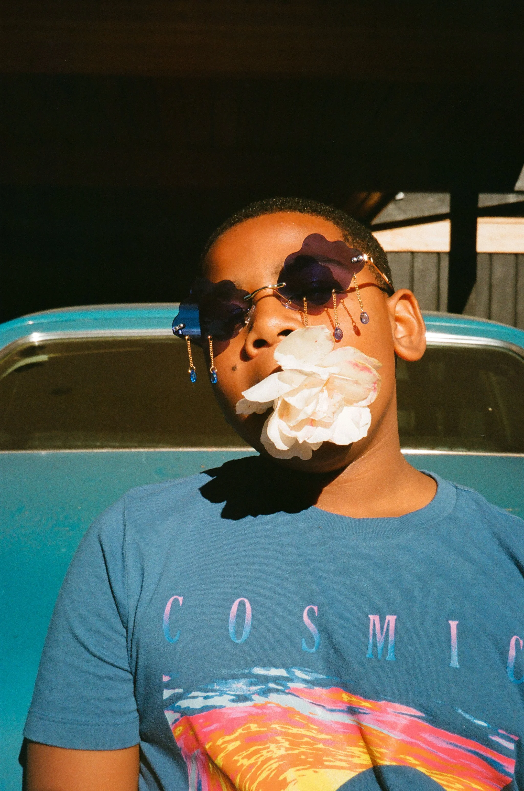 A boy wearing dark sunglasses with flower decorations, holding a white flower in his mouth, standing in front of a vintage nova teal car . Film photography Tacoma Washington