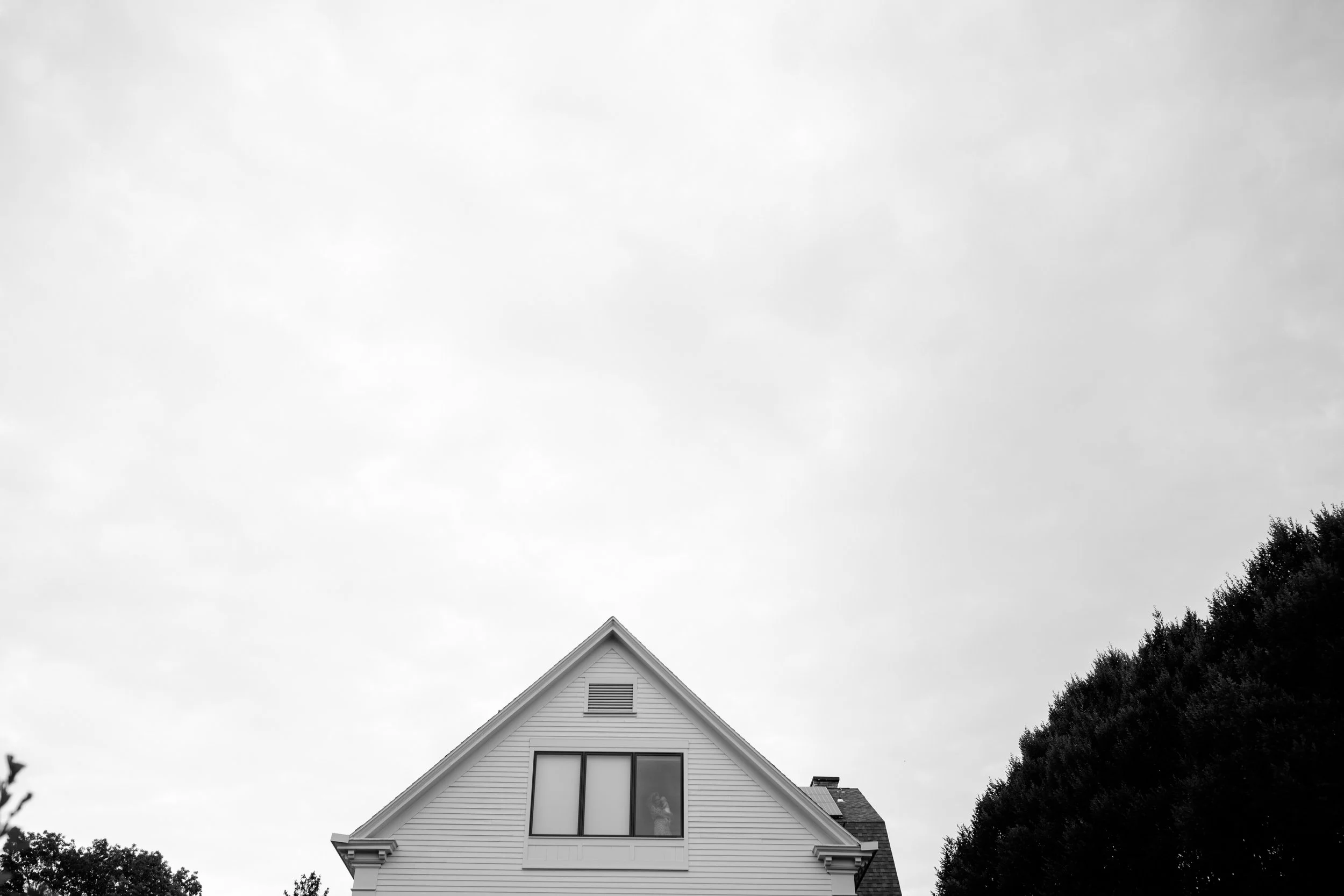 A black and white photo of a house with a gabled roof, centered in the frame, with trees on either side and a cloudy sky above.