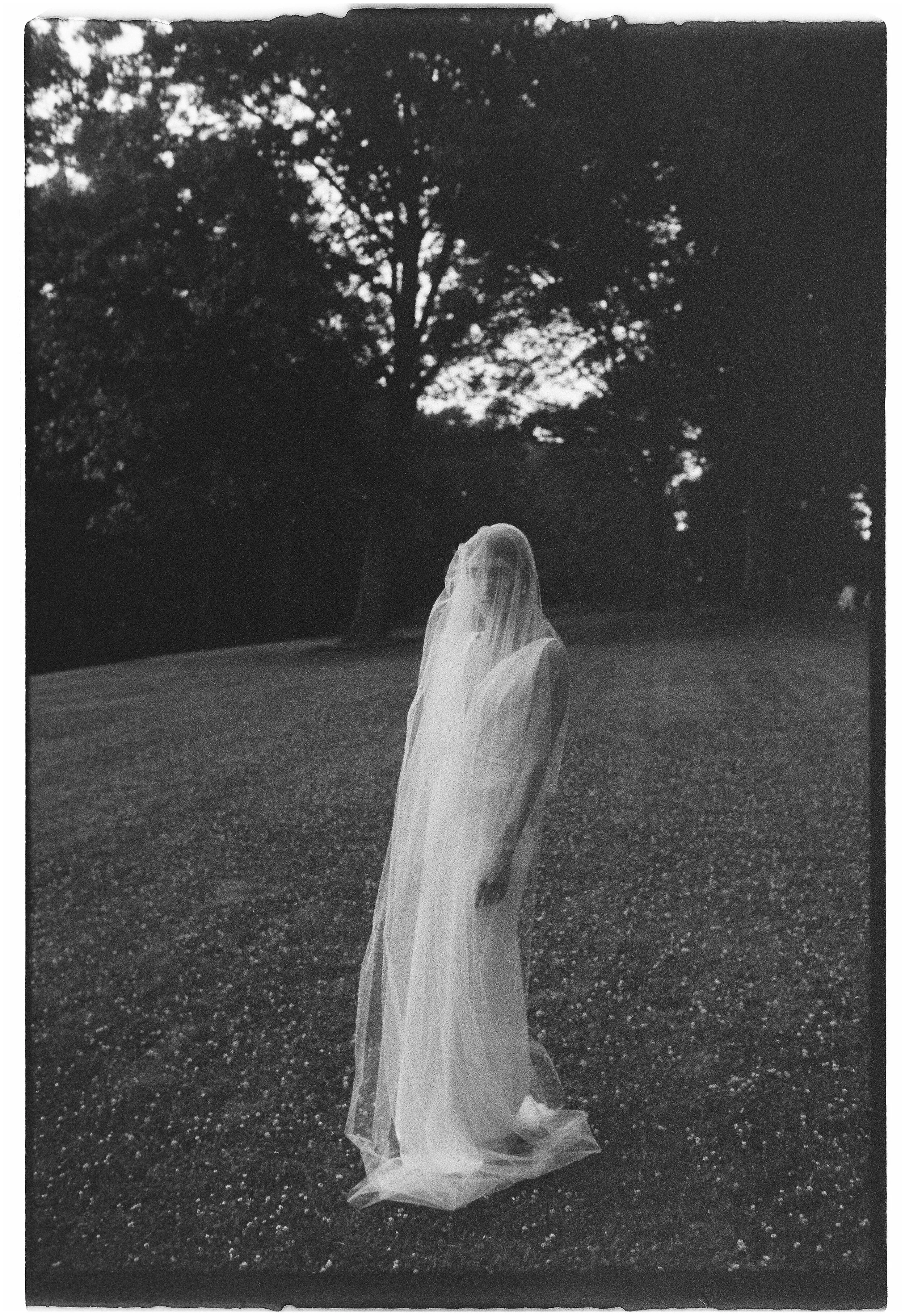 Black and white film photograph of a bride wearing a wedding dress with a theatric veil. Alone in a field.