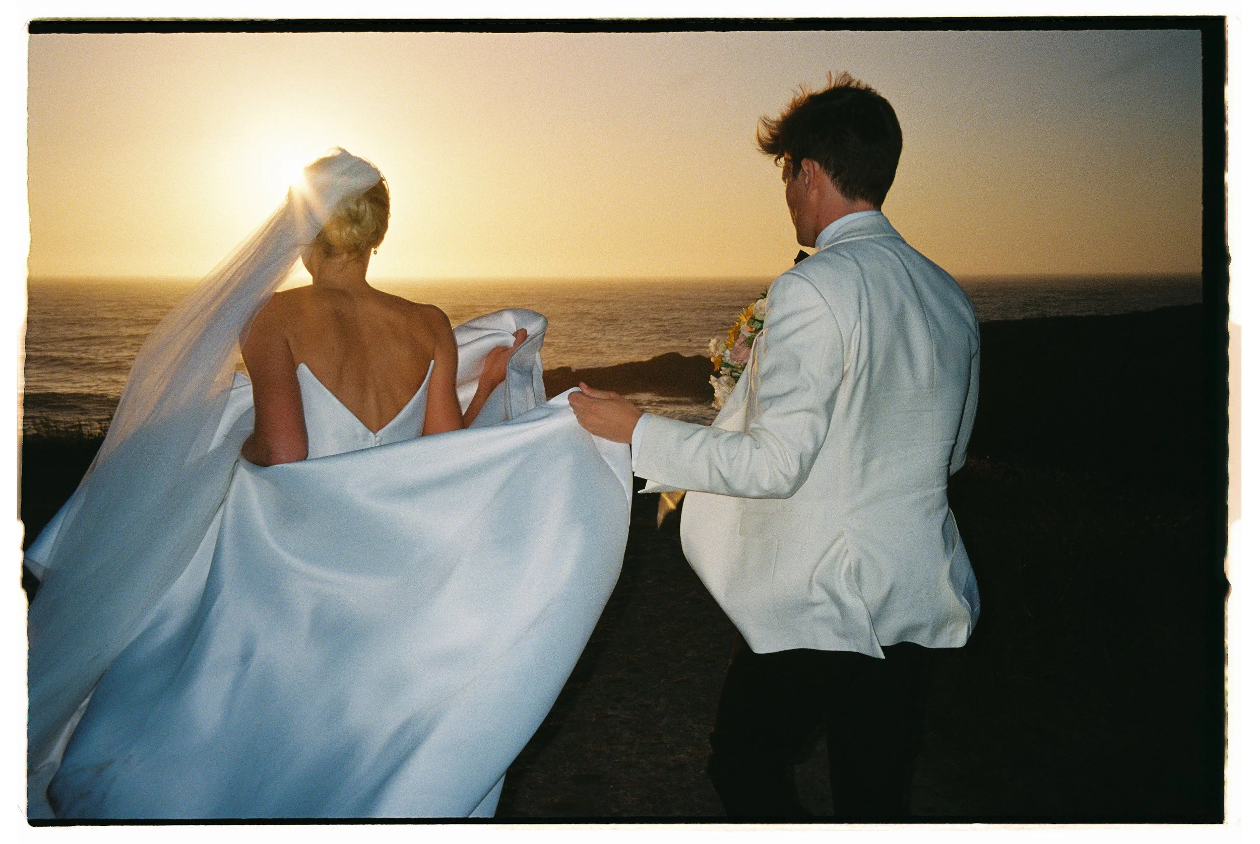 A bride and groom at a beach sunset, with the bride in a strapless wedding dress and veil, and the groom in a white tuxedo, kneeling and holding the bride's dress. Film, Heritage house, Mendocino, California. The highlands. 