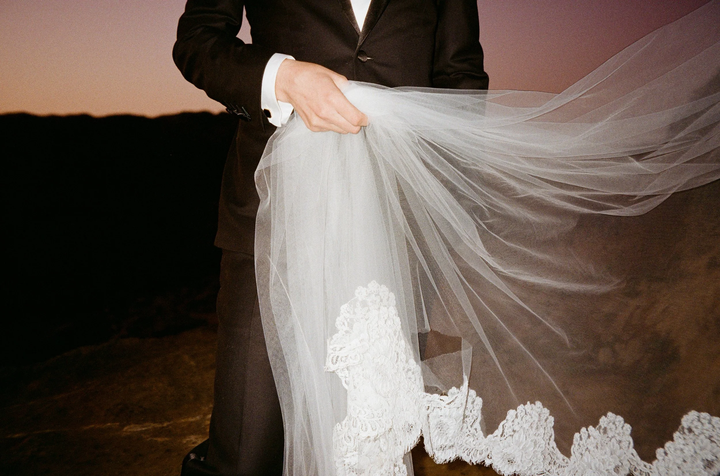 A groom in a black suit holding the train of a wedding dress with lace detailing at sunset or dusk.