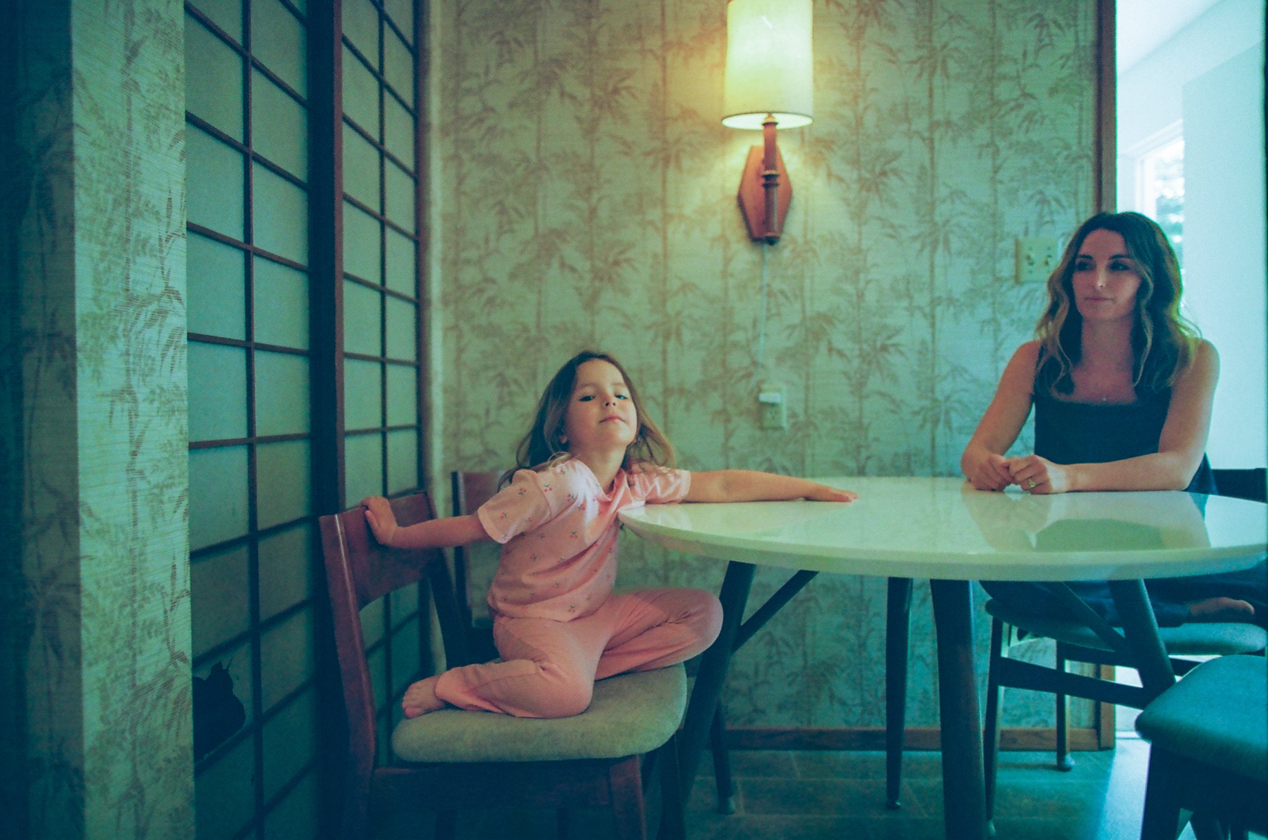 A young girl in pink pajamas is sitting on a chair at a dining table, stretching her arms and legs, with an adult woman sitting across from her, smiling.