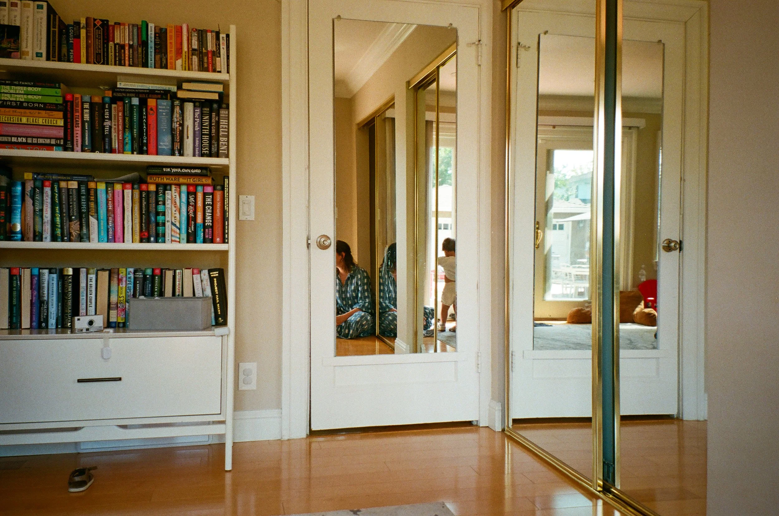 View of a room with a white bookshelf filled with books on the left, and for-albeit mirrored closet doors reflecting a seated woman in pajamas, a child standing, and a view of a backyard with a deck and sliding glass door. Film photos