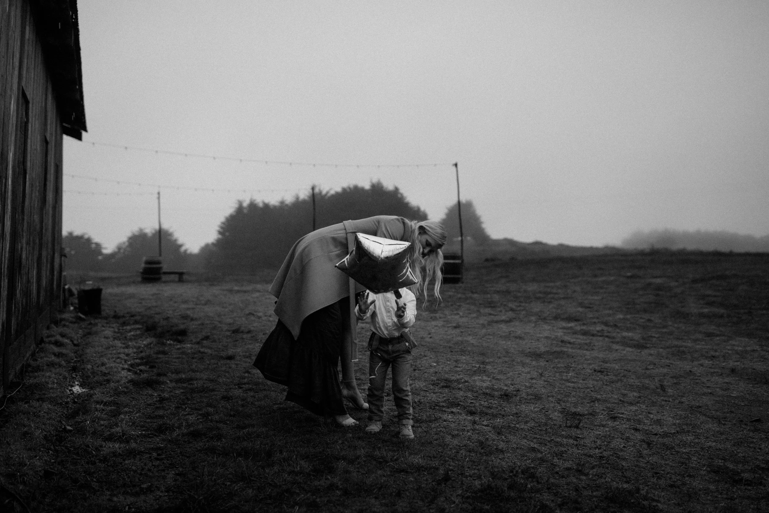 A woman and a child with a balloon on a farm, standing beside a wooden barn, with trees and a swing set in the background, in black and white.