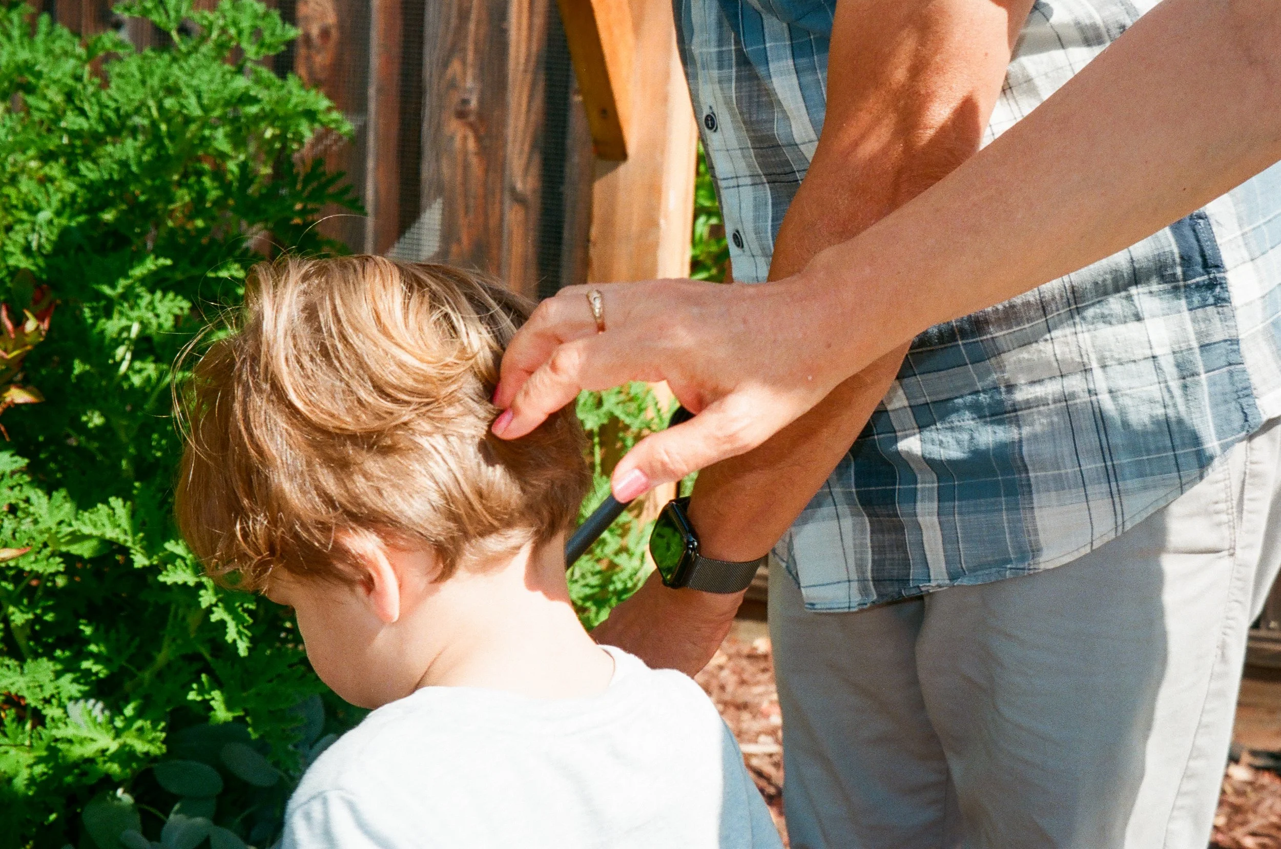 An adult adjusting a young boy's hair outside during daytime, with a wooden fence and green plants in the background.
