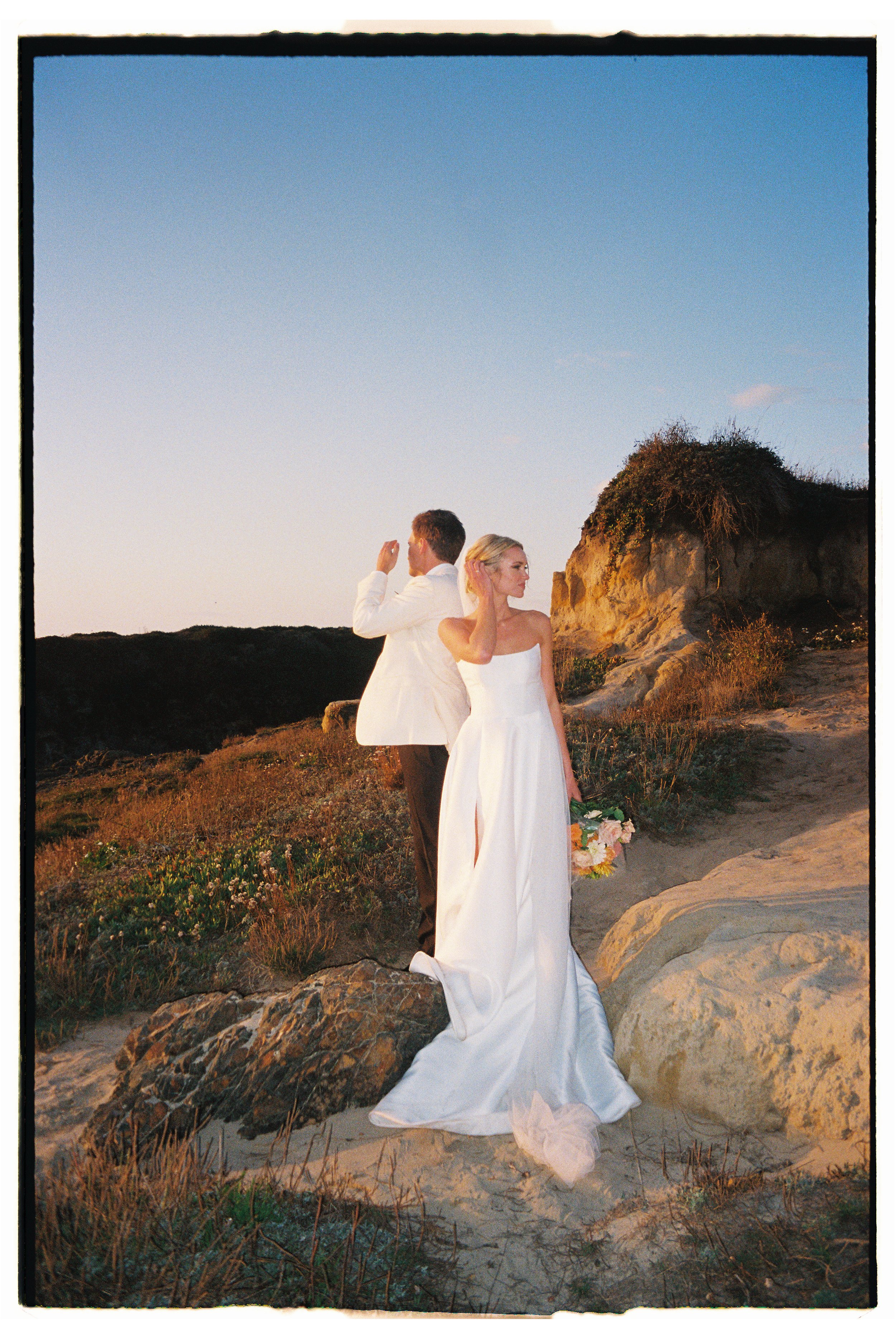 A bride and groom standing on a rocky outdoor landscape at sunset, with the bride holding a bouquet and the groom wearing a white jacket and black pants.