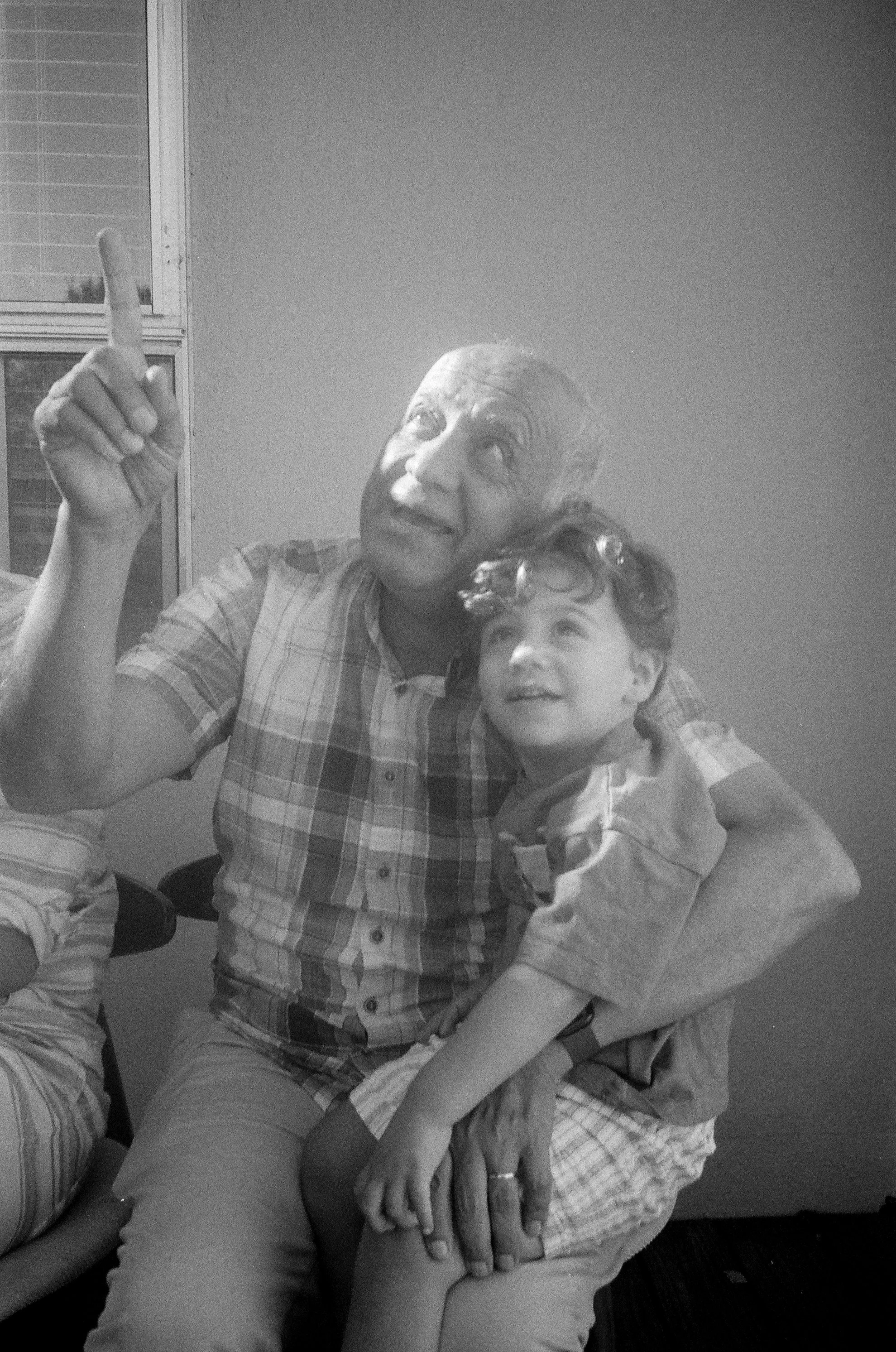 An older man with glasses pointing upward, sitting with a young boy on his lap, both looking upward with interest. Palo Alto family photos in film. 