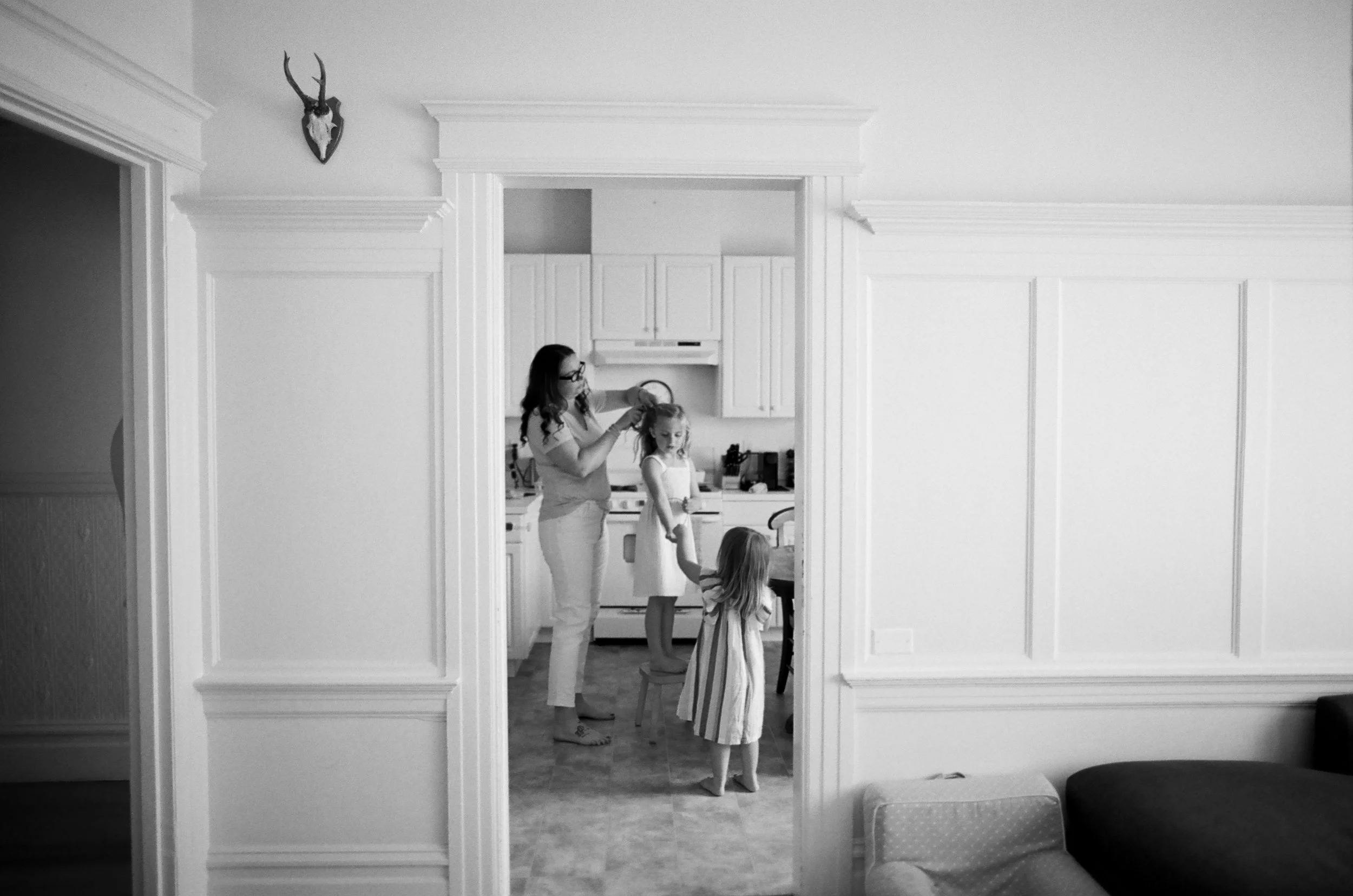 A woman with glasses and two young girls in a kitchen, with one girl sitting on a chair and the other standing, as the woman styles the sitting girl's hair.