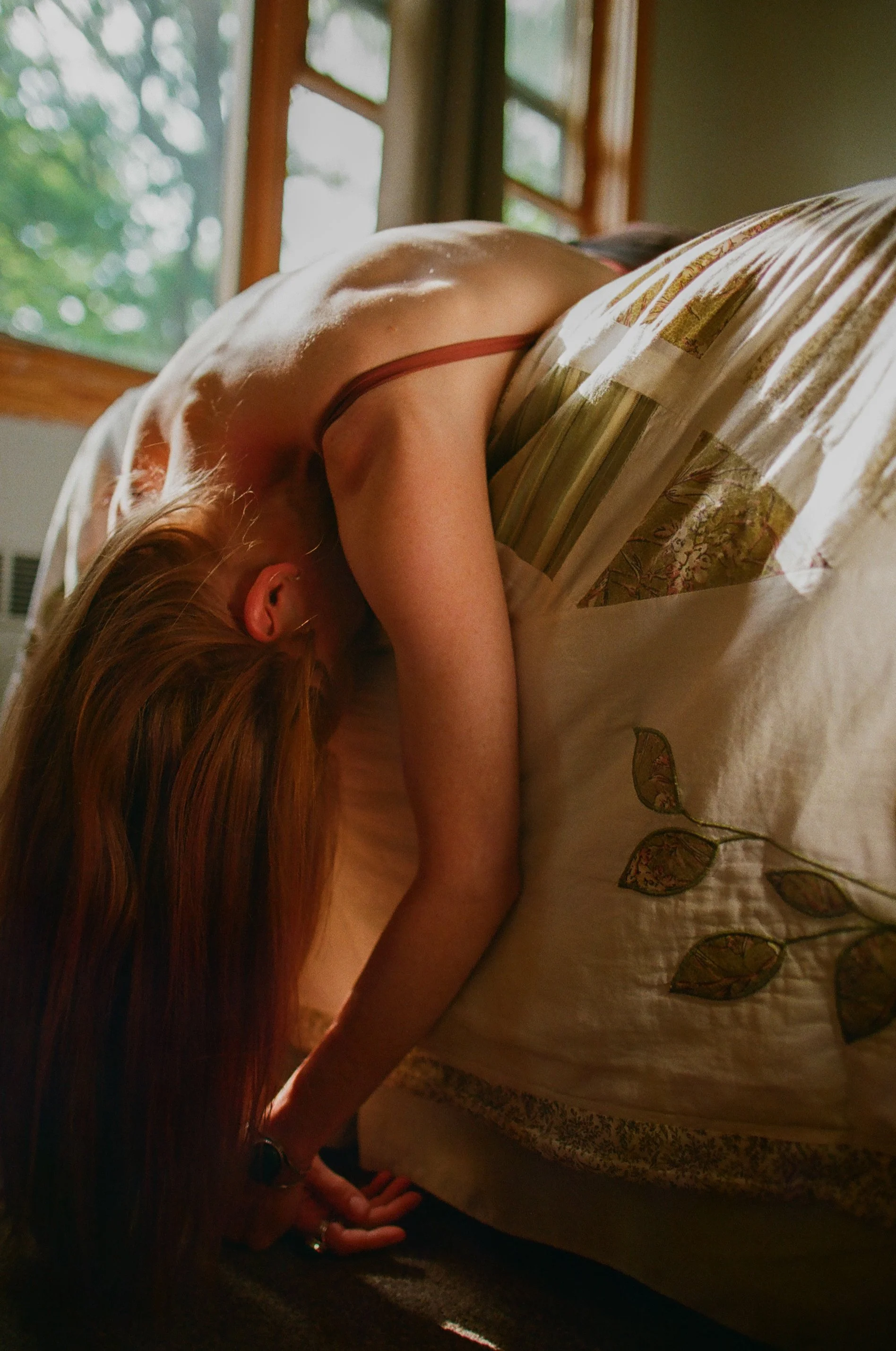 A woman with red hair leaning forward over the edge of her bed, with her head bent down and hair covering her face. She is wearing a thin strap top, and the bed has a cream-colored blanket with leaf embroidery. The room has a wooden window with green