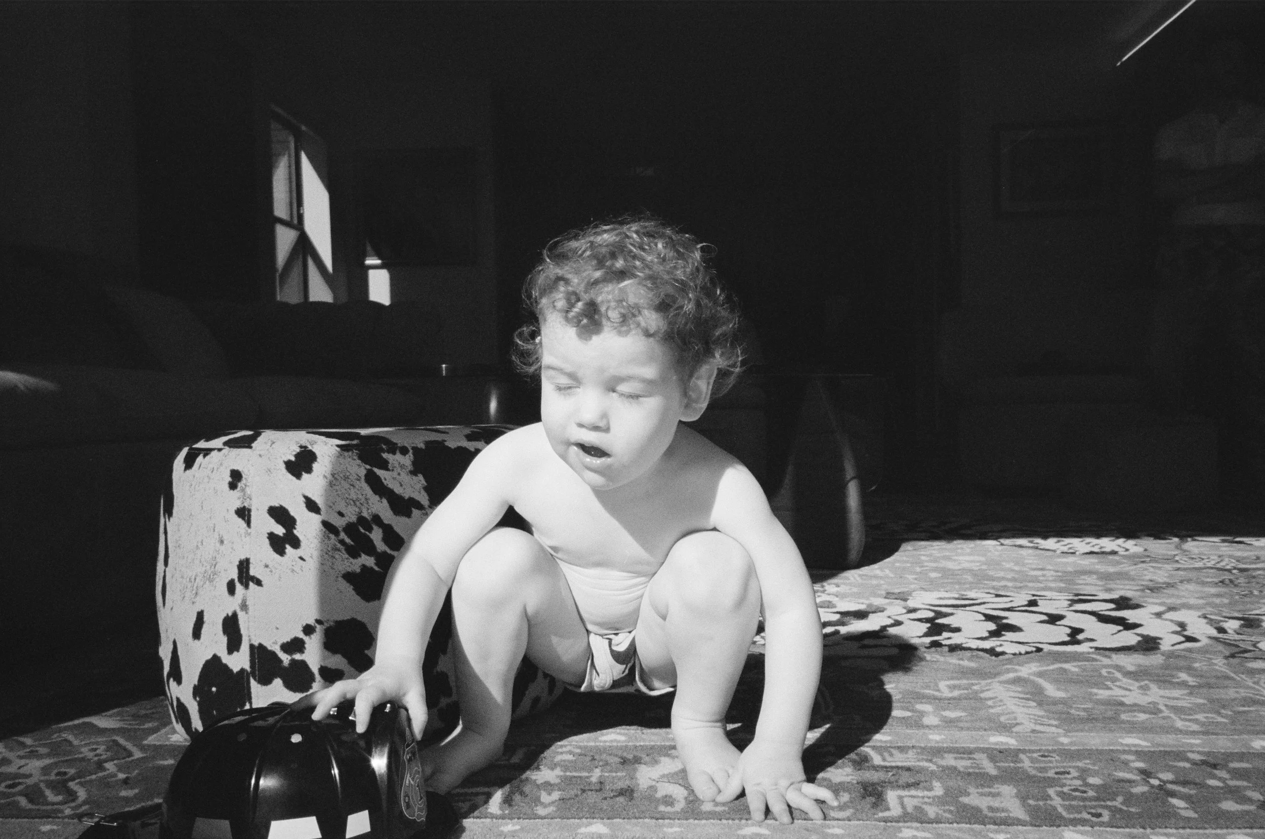 A baby with curly hair wearing a diaper, sitting on a patterned rug inside a room. The baby is reaching out to touch a black toy with a face on it, nearby a leopard-print ottoman.