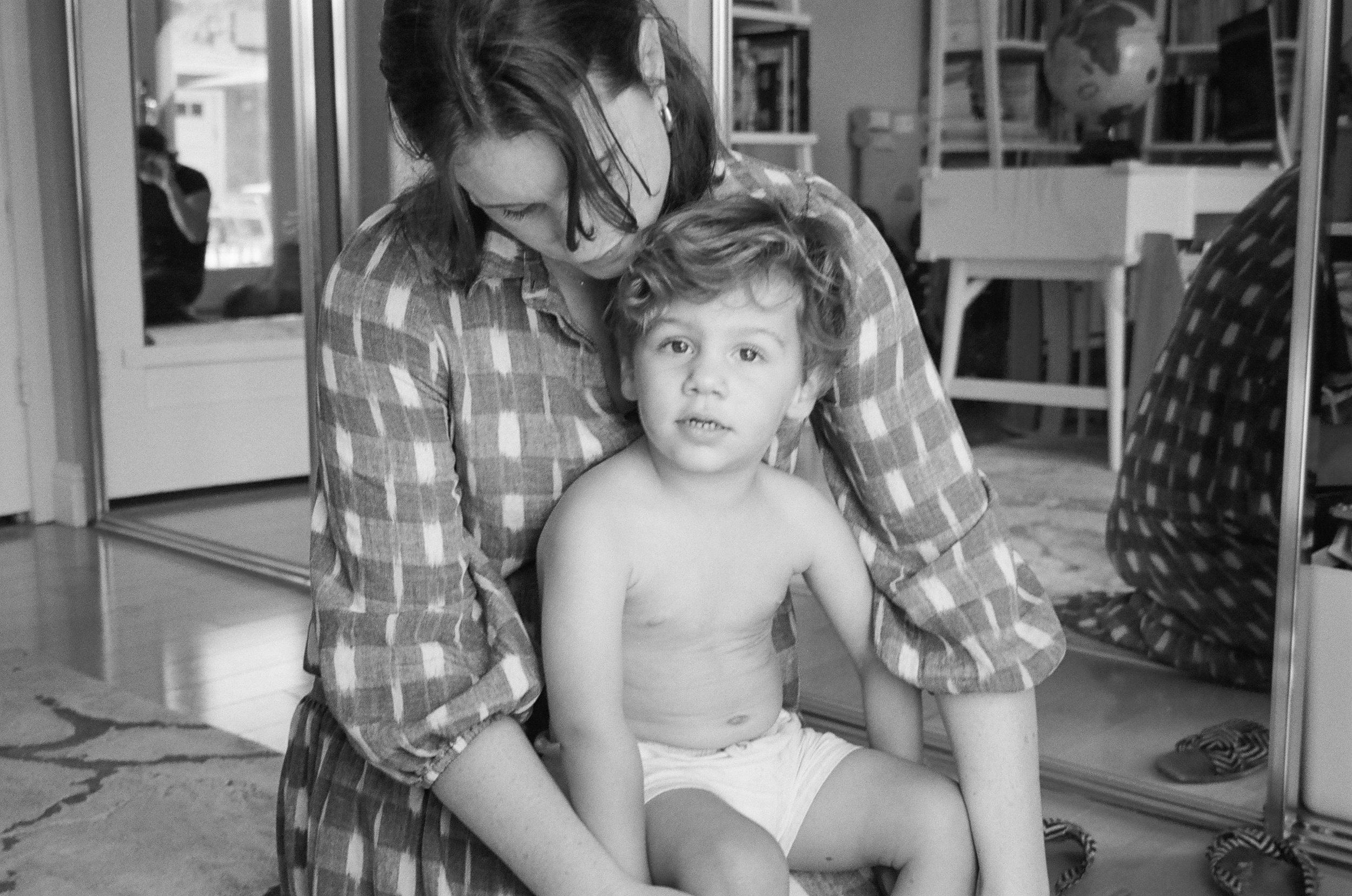 A woman with shoulder-length hair, wearing a plaid shirt, crouches down and embraces a shirtless young boy with curly hair sitting on her lap inside a room. In the background, a person is seen through a glass door taking a photo.