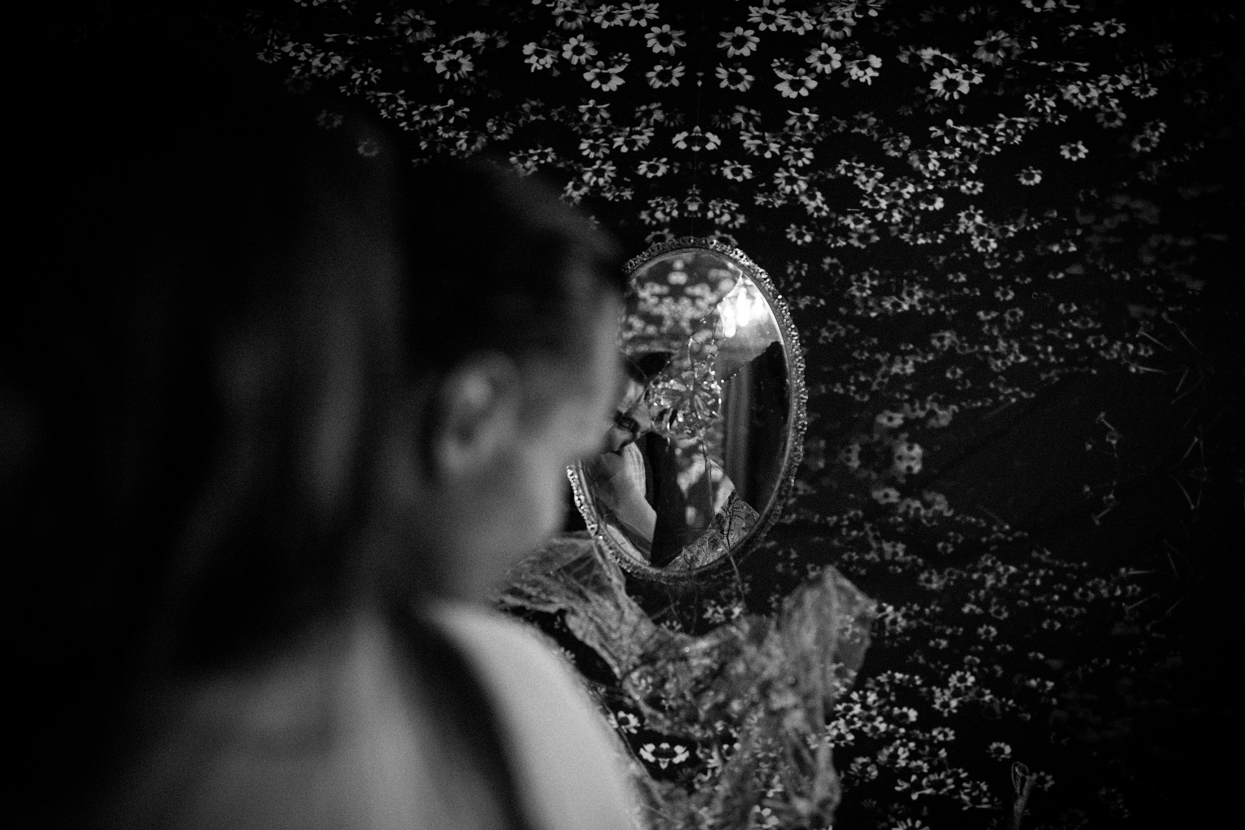 A woman looks at her reflection in a small, oval mirror with a decorative frame. She is surrounded by a floral-patterned cloth or curtain, and her face is partially visible, in profile.