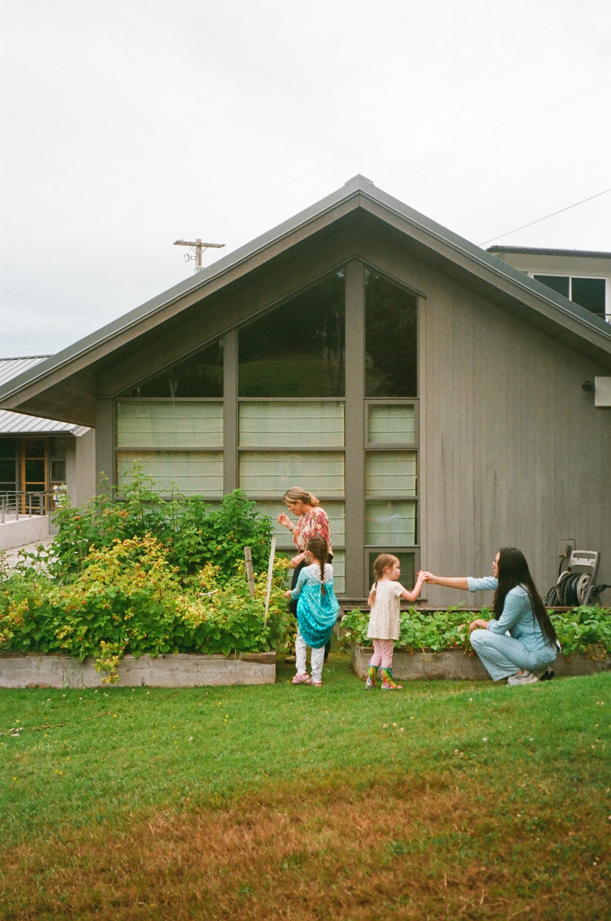A woman and four children in a garden outside a modern house with large windows, the woman is squatting and the children are standing, two of them are holding hands and one of the children is wearing a superhero cape.