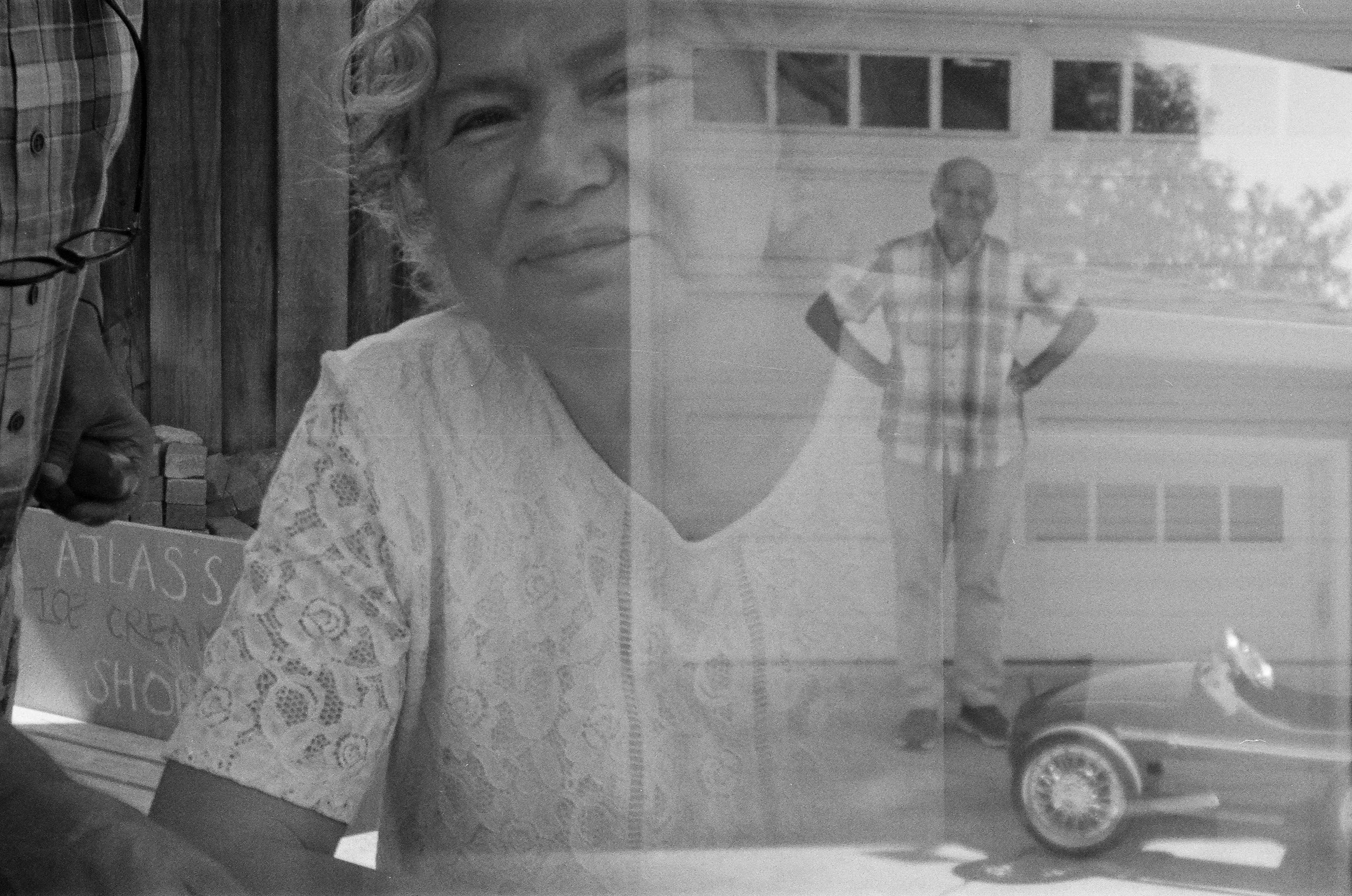 A woman and behind her, a man stands with his hands on his hips, wearing glasses and a plaid shirt, outside in front of a house and a car. Multi generational double exposure film photo Palo Alto, California family photos. 