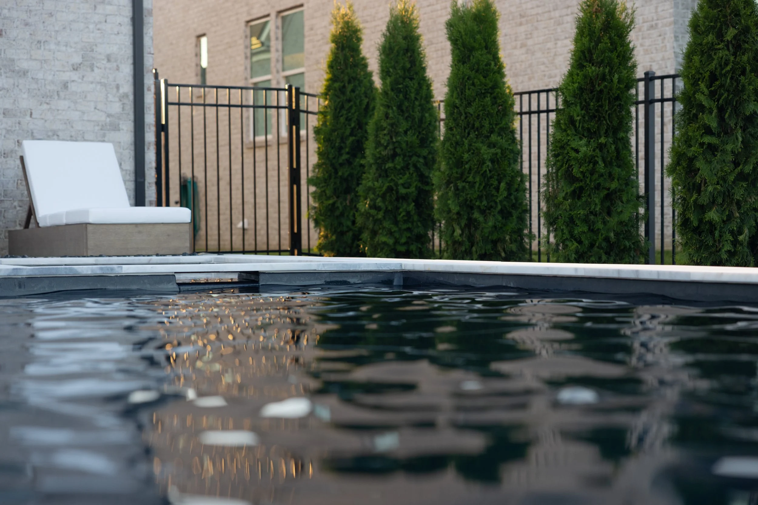 Close-up of a swimming pool's water edge with reflections on the surface, a white lounge chair on a wooden platform, a black metal fence, tall green trees, and a beige brick building in the background.