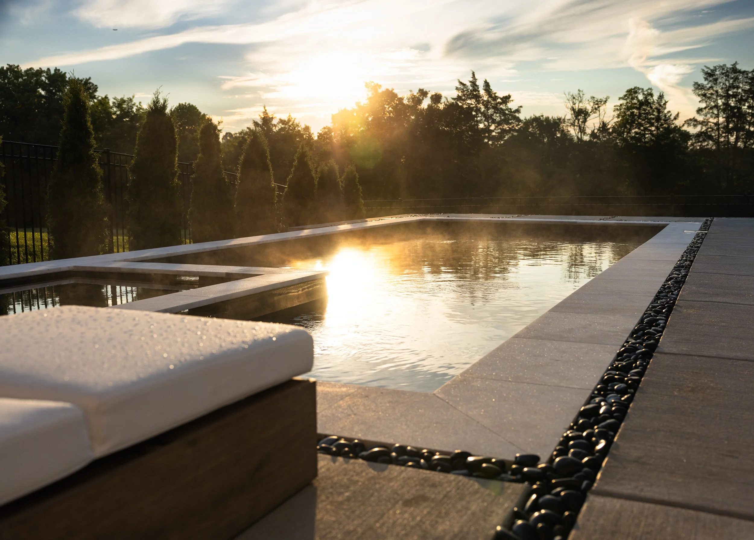A pool with steam rising, surrounded by a marble deck with a decorative border of black stones, overlooking a landscape with trees and a sunset in the background.