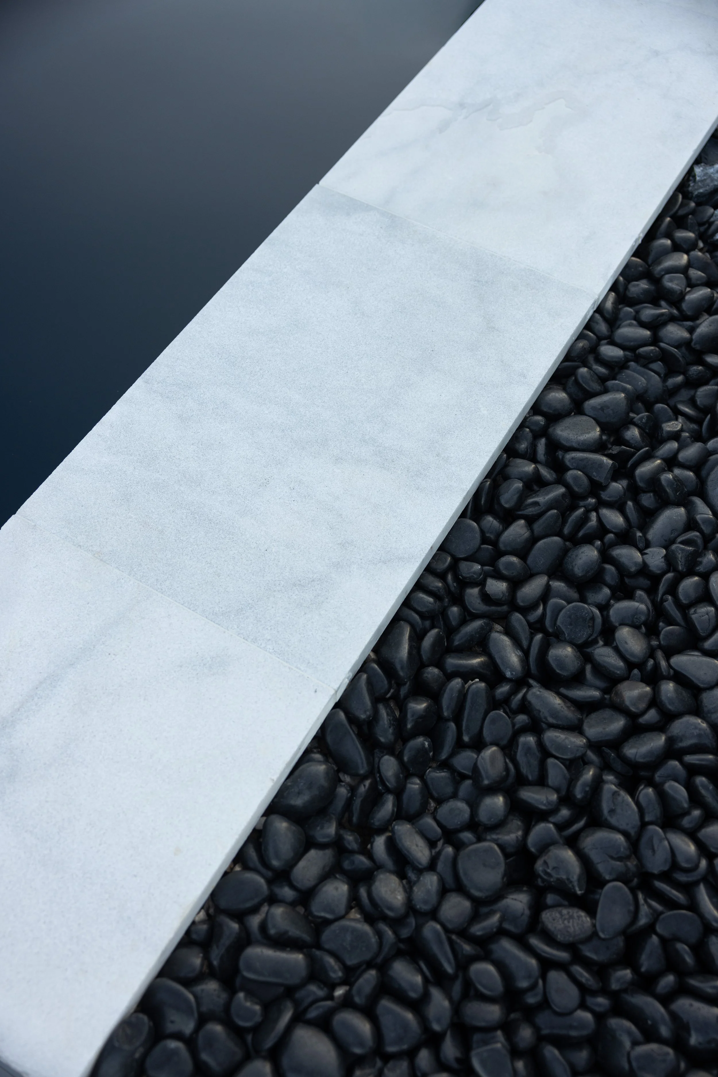 A close-up of a modern aspect of a swimming pool with a white stone edge, black smooth pebbles surrounding it, and the water reflecting the sky.