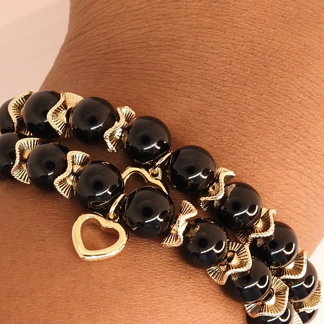 A close-up of a bracelet made of black beads, golden decorative elements, and a gold heart-shaped charm on a person's wrist.