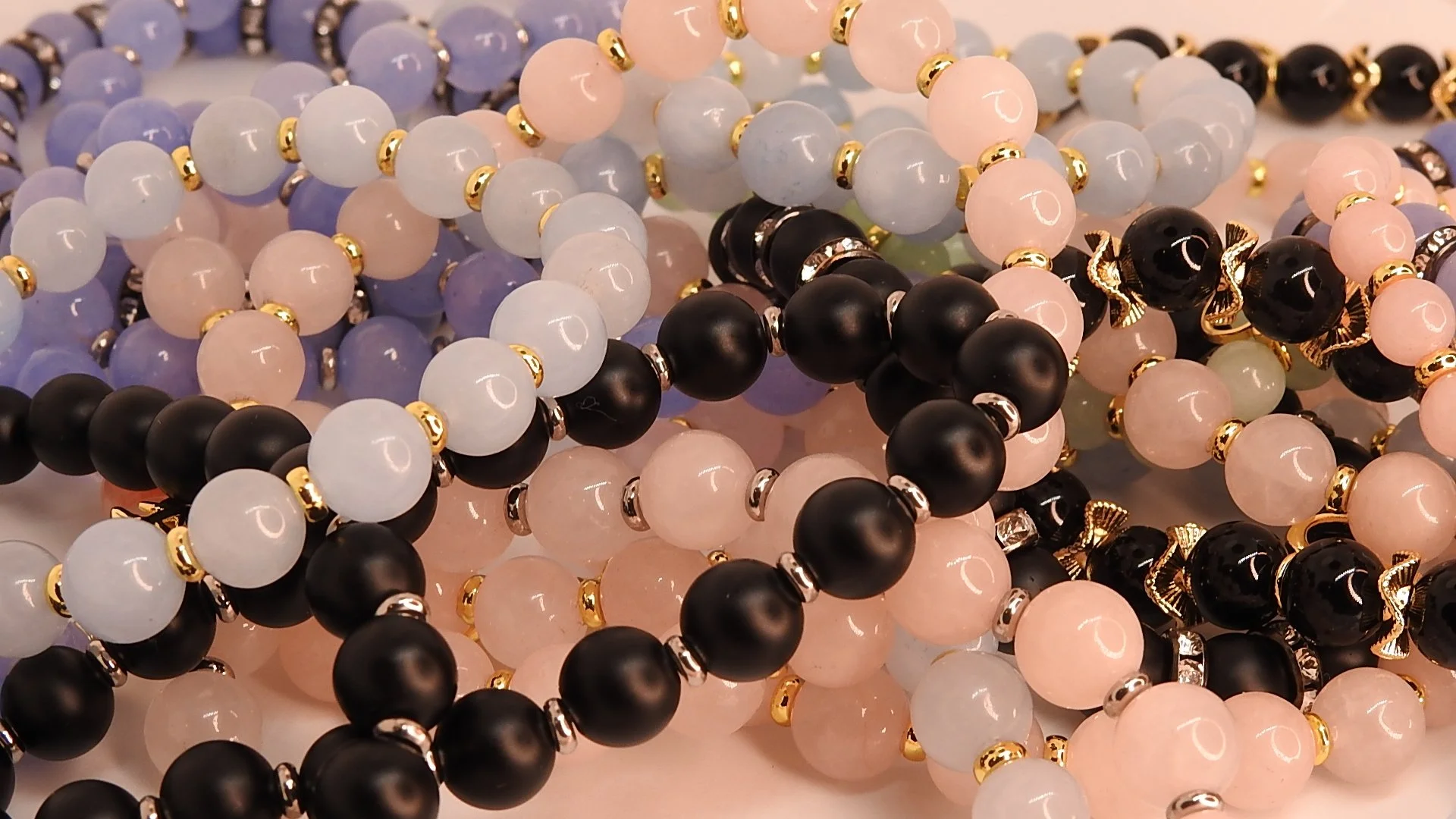 Close-up of multiple beaded necklaces in black, pink, light blue, and purple, with some having gold accents, arranged on a flat surface.
