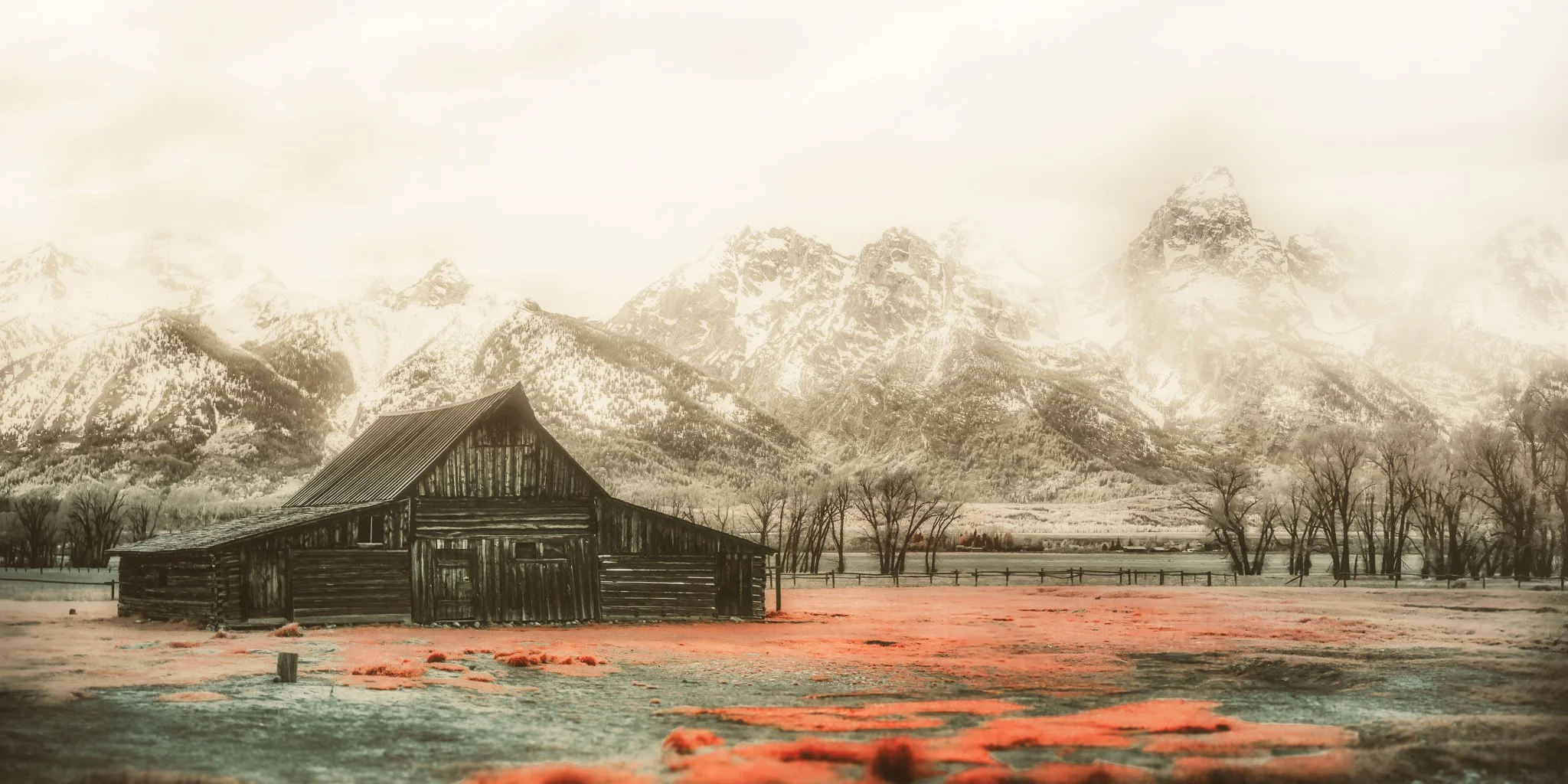 A rustic wooden barn in a field with mountain range in the background, trees, and a fence, captured in sepia tone.