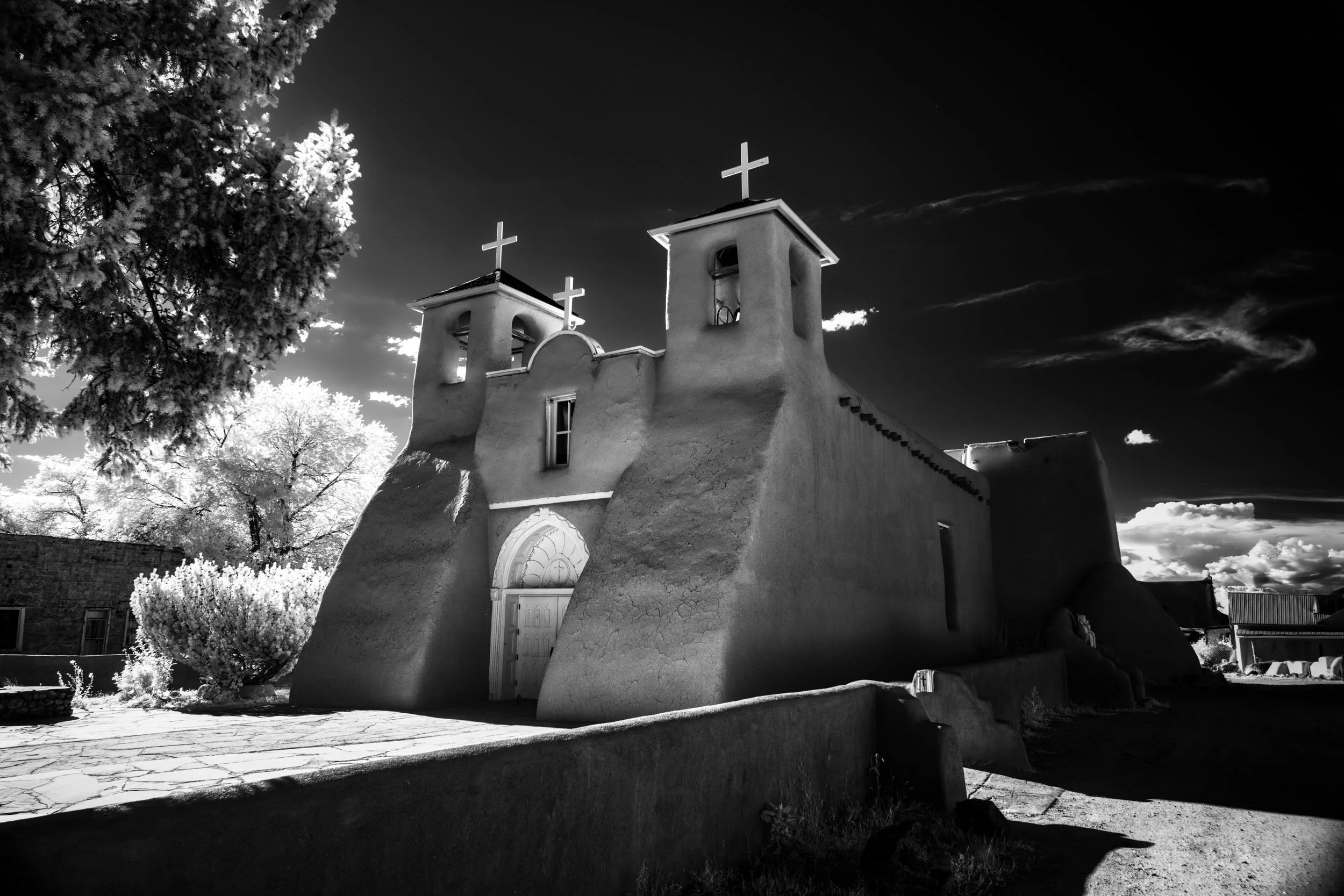 Sanctuary of Timeless Shadows — The historic adobe church of San Francisco de Asís rises in infrared black and white, its twin bell towers against a dramatic sky.