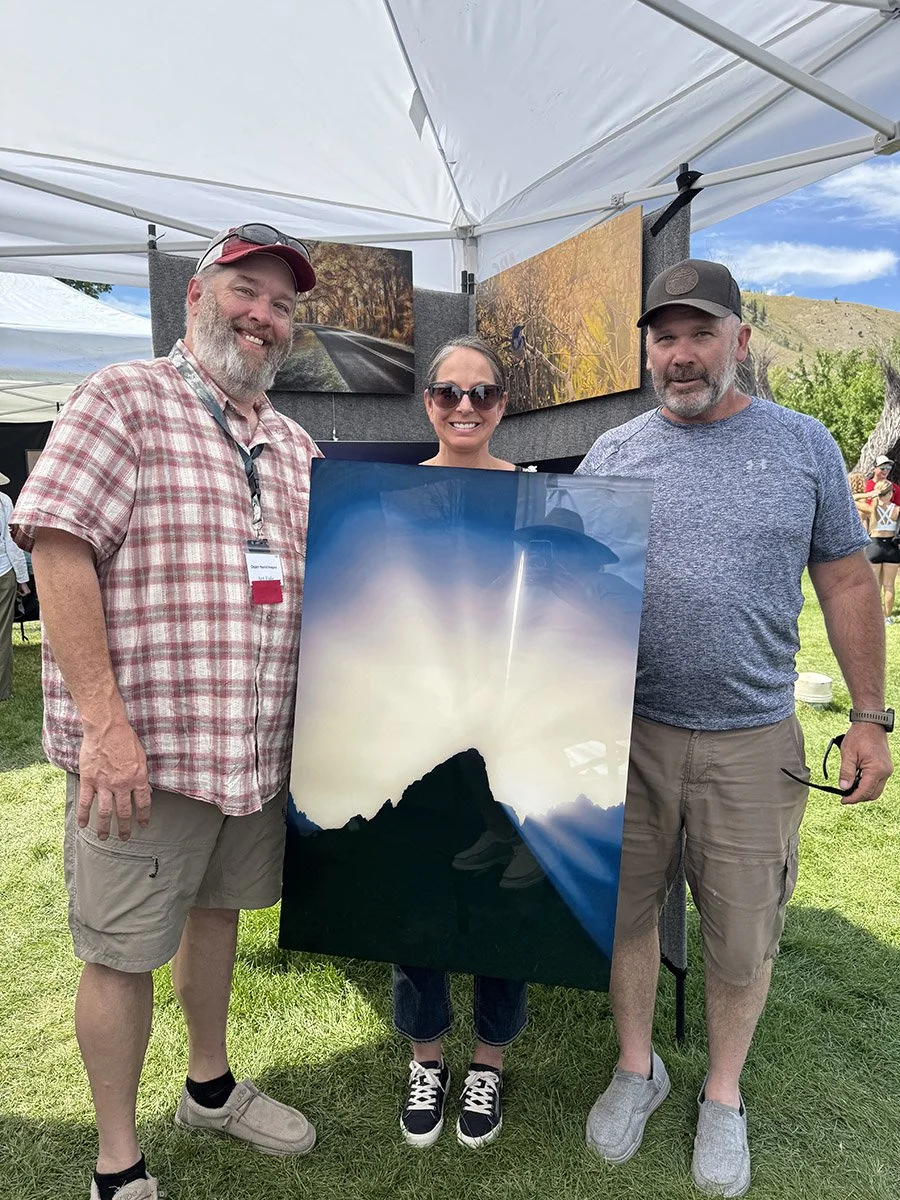 Three people standing outdoors under a white canopy, holding a large landscape photograph featuring mountains, trees, and sunlight, at an art or craft fair.
