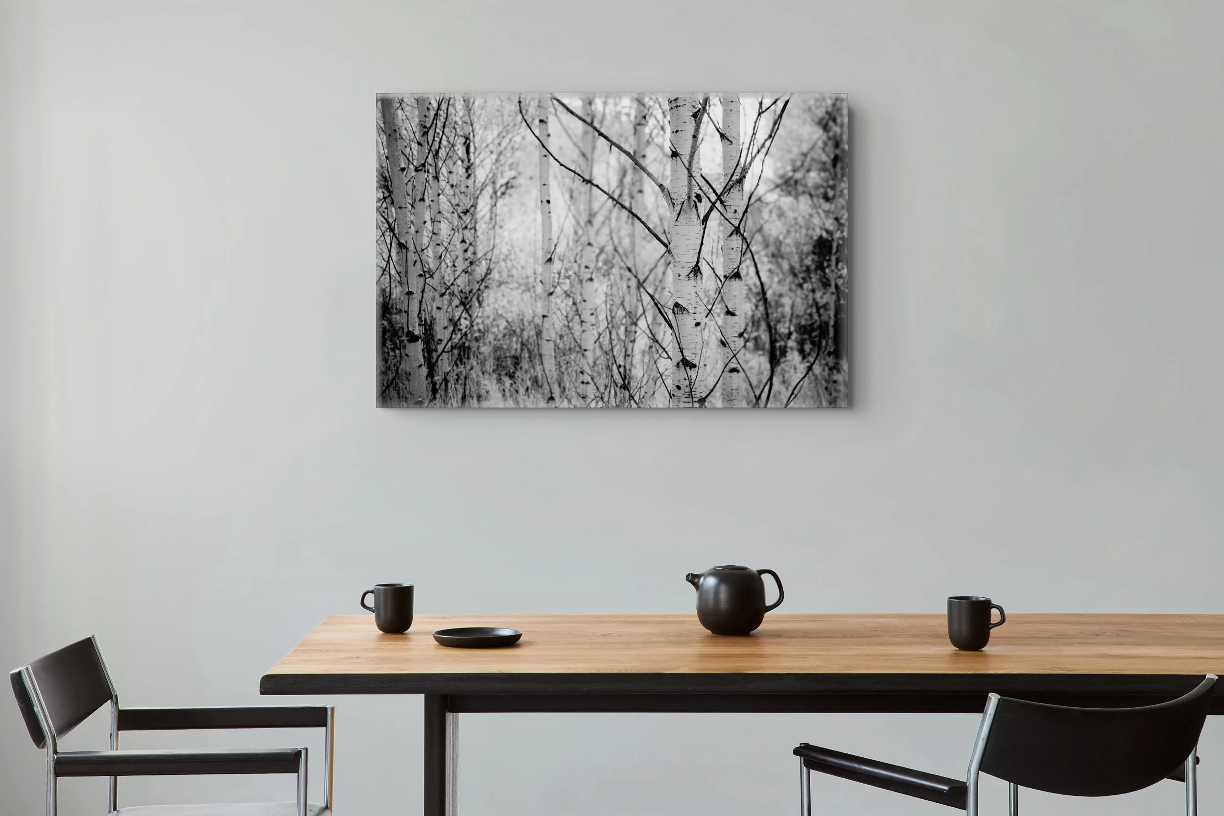 Black-and-white photograph Contrasts in Woodland Quiet displayed above a wooden dining table in a minimalist room with gray walls and black chairs.