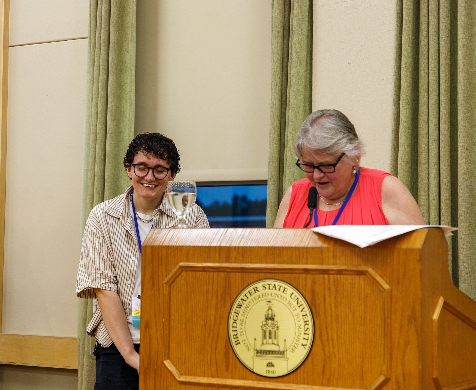 A young man and an older woman are standing at a podium at a university event. The woman is reading or speaking into a microphone, and the young man is smiling. There is a glass of water on the podium and green curtains in the background.