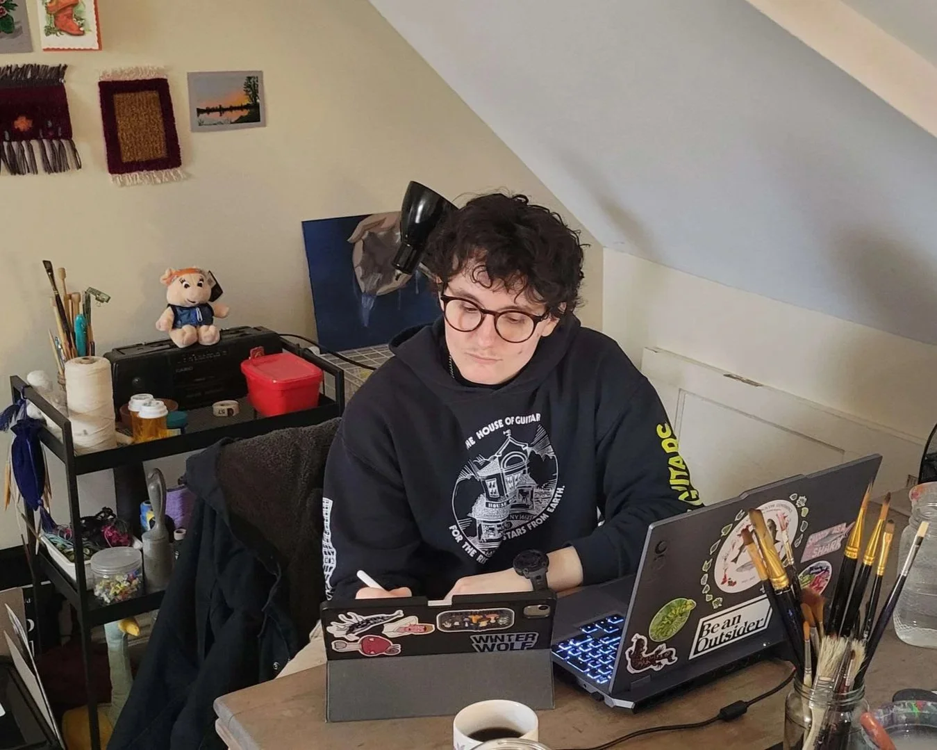 A person with curly dark hair and glasses working at a desk with a laptop decorated with stickers. The person wears a black hoodie with designs related to guitars. The workspace has art supplies, a shelf with art materials and a plush toy, and a cup 