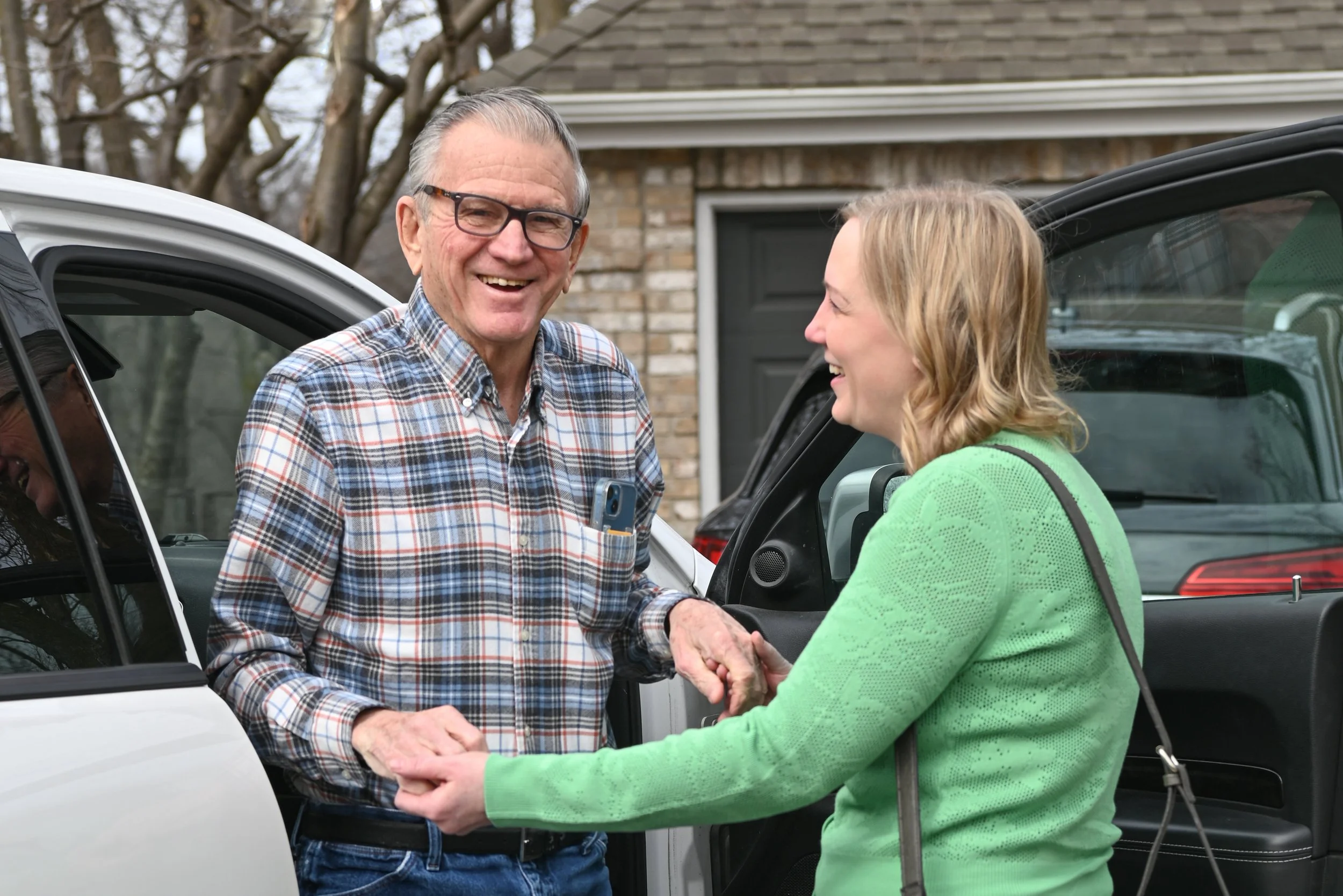 A cheerful elderly man and a smiling woman are shaking hands outside near parked cars, with a house in the background.