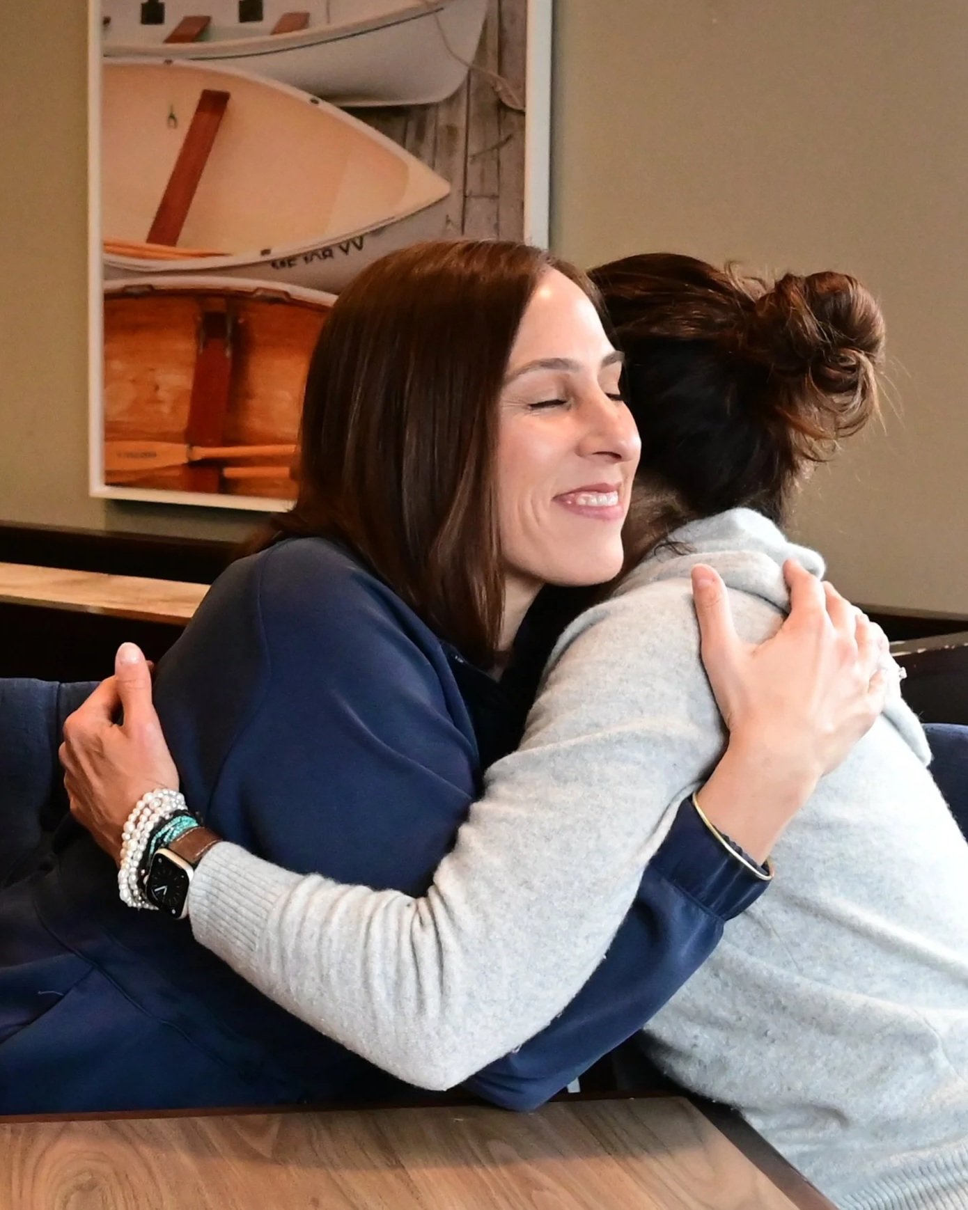 Two women hugging and smiling in a warm, friendly manner at a table in a cozy setting.